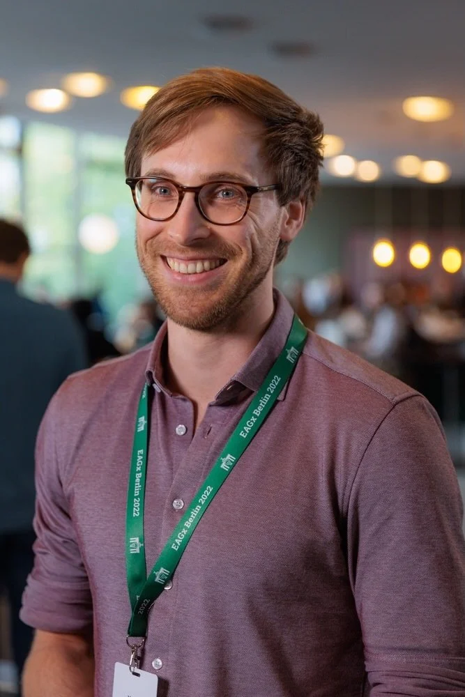 Man smiling wearing glasses and a green lanyard, indoor event setting, blurred background with lights.