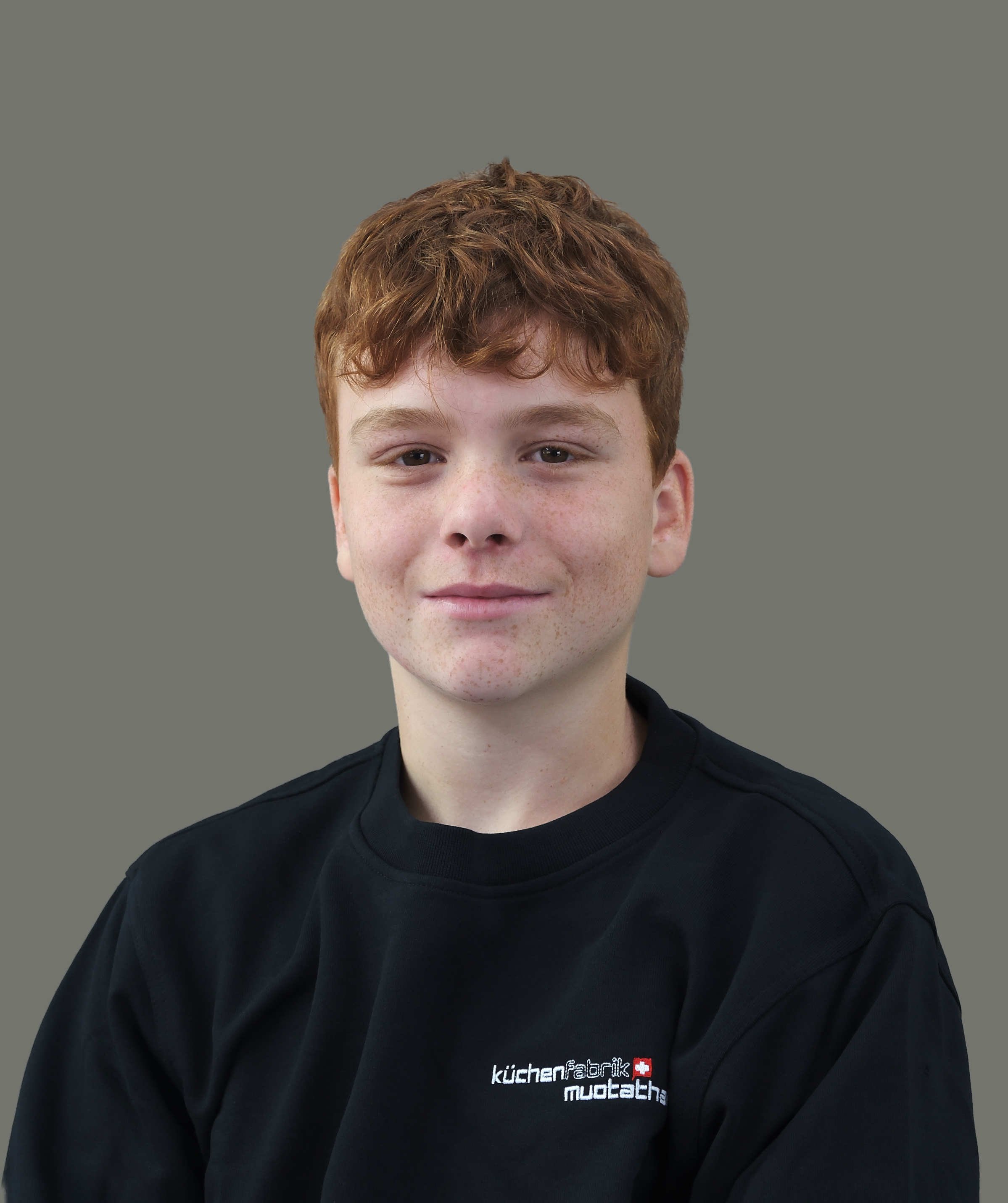 Junge mit roten Haaren, trägt schwarzen Pullover mit weißen und roten Schriftzug 'küchenfabrik nuotha' auf schwarzem Hintergrund, vor grauem Hintergrund.