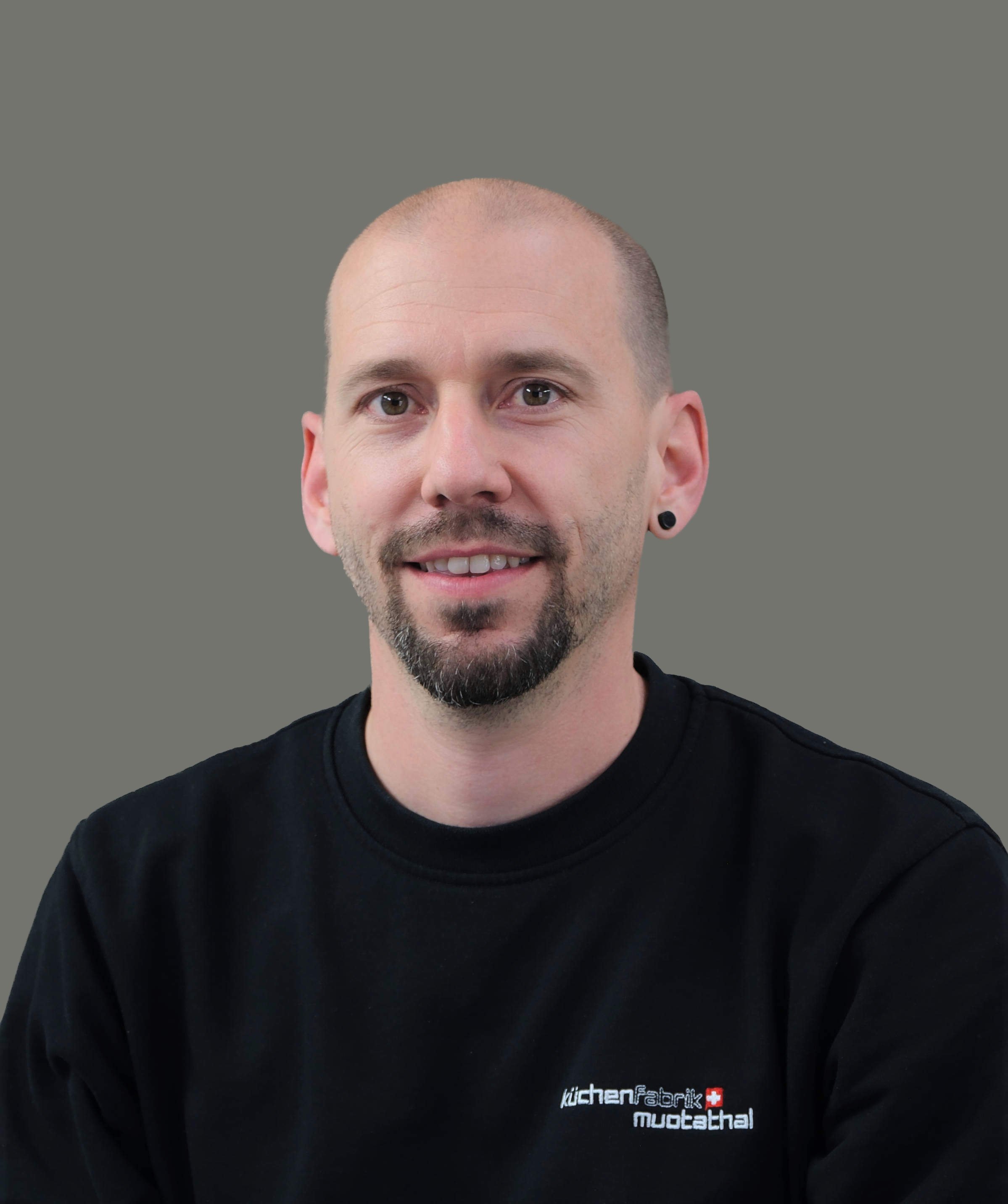 Nahaufnahme eines Mannes mit Kurzhaarschnitt, Brauen, dunklem Bart und schwarzen Ohrringen, trägt ein schwarzes T-Shirt mit weißem und rotem Text.