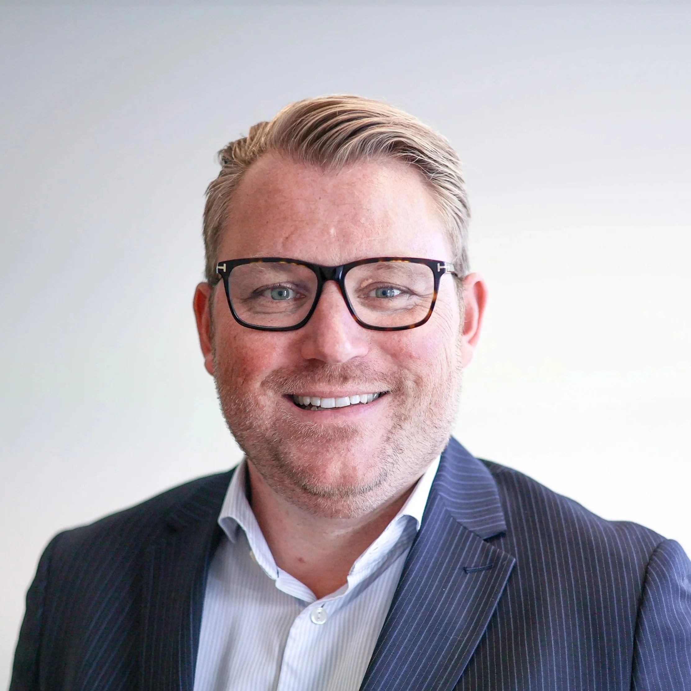 A man with blonde hair, wearing black-rimmed glasses, a navy blue pinstripe suit, and a white shirt, smiling at the camera against a plain light background.