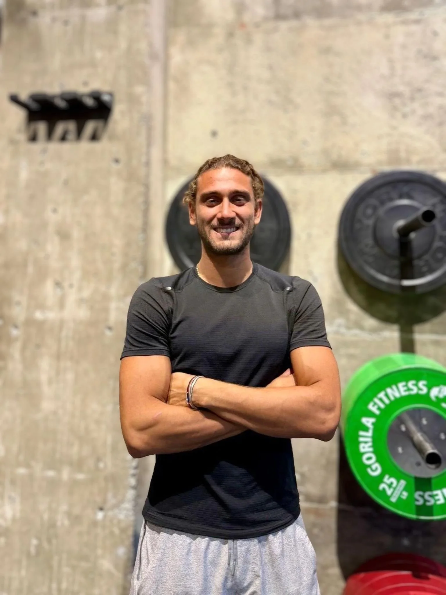A man smiling with arms crossed in a gym, standing against a concrete wall with weightlifting plates, including a green Gorilla Fitness weight plate and black plates, behind him.