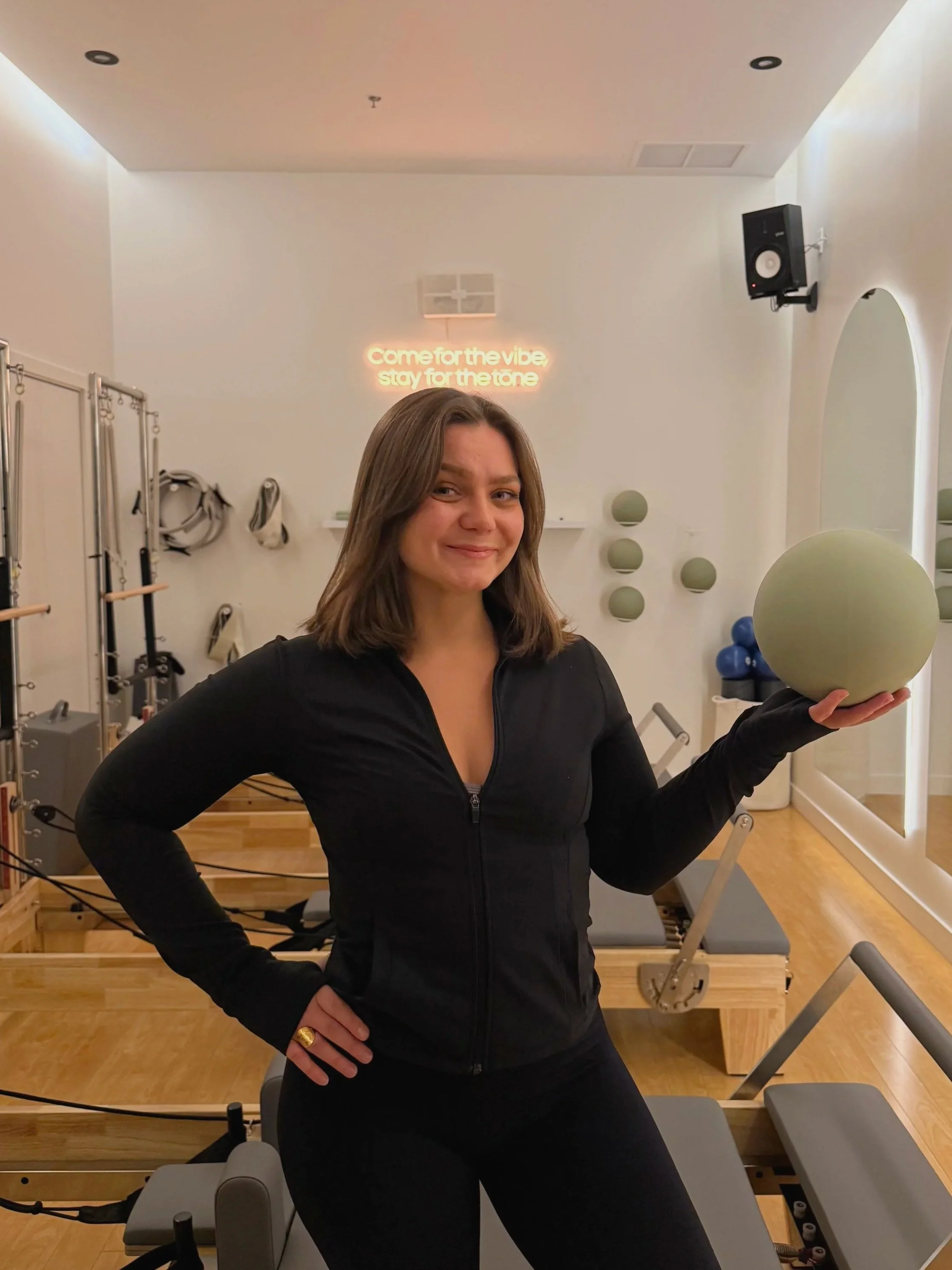 A woman in a black outfit holds a green exercise ball in a modern Pilates studio