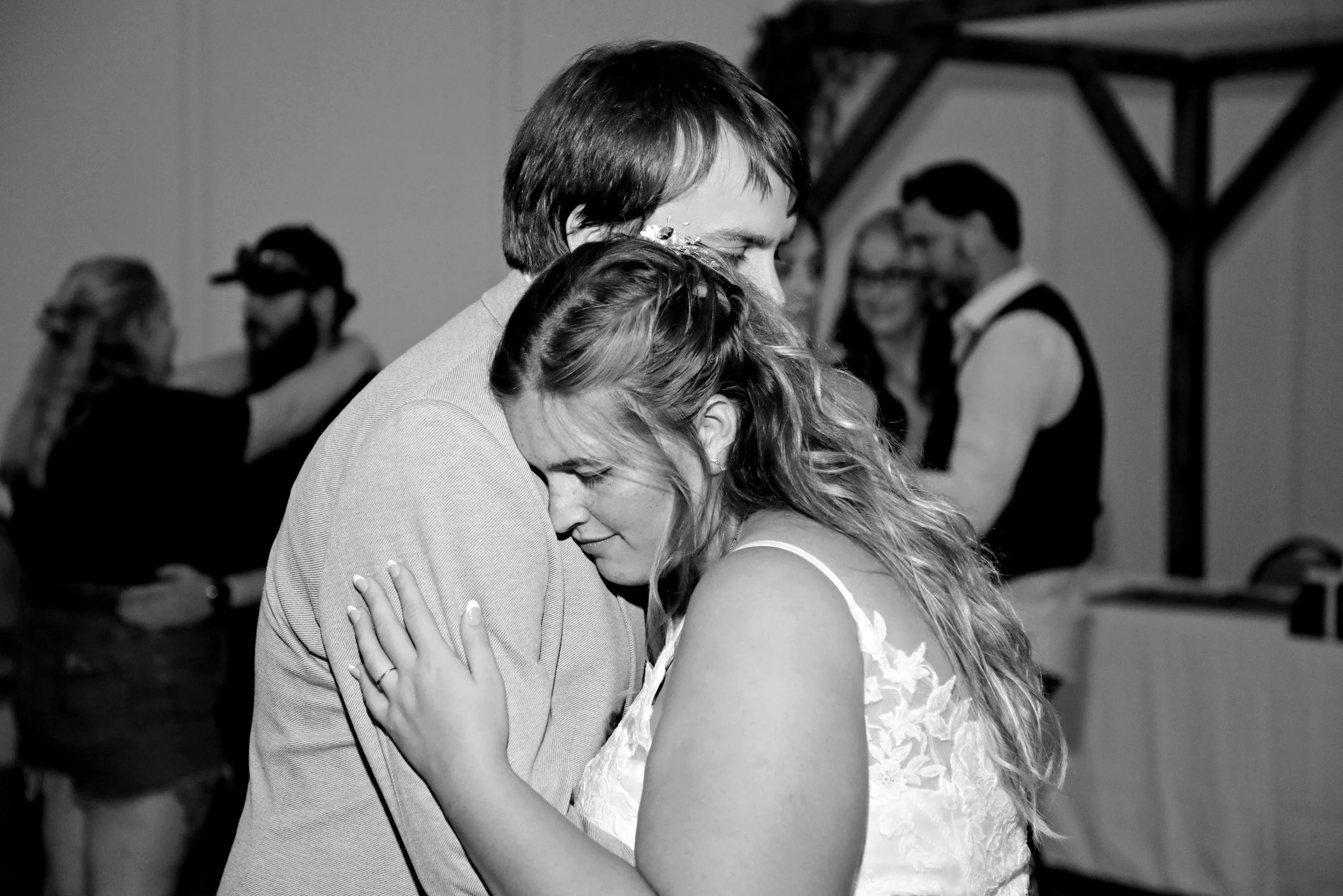 A black and white photo of a couple embracing during a dance at their wedding, with other people in the background also dancing and celebrating.