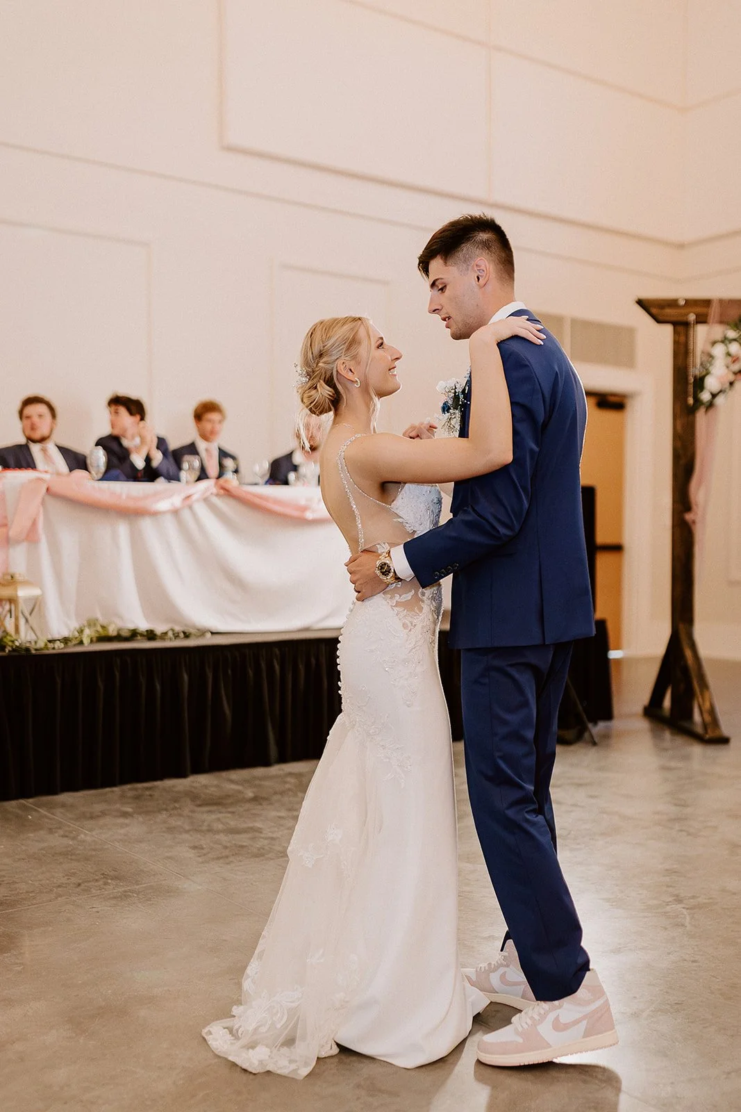 A bride and groom dance at their wedding reception, with the bride in a lace wedding gown and the groom in a blue suit and white sneakers.