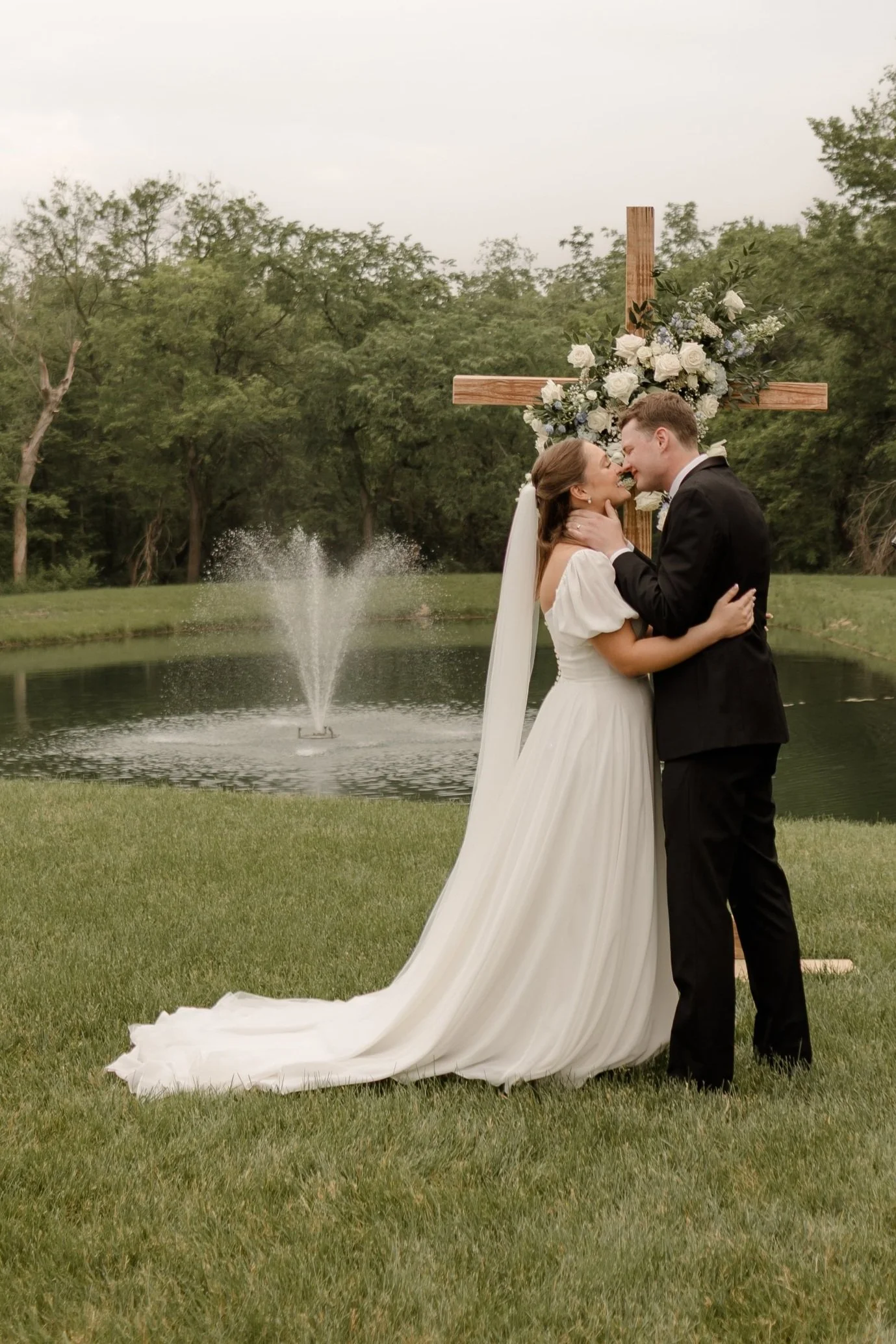 Outdoor wedding ceremony at Northbridge Event Center with bride and groom sharing their first kiss in front of a floral cross, surrounded by guests and scenic greenery—a joyful moment full of love and celebration in central Iowa.