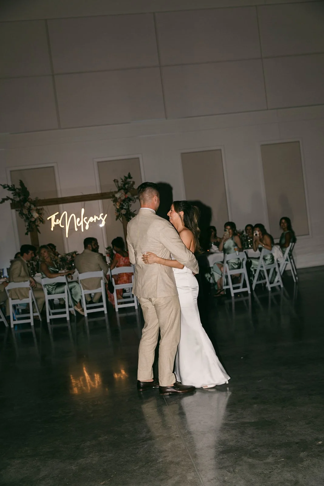 A bride and groom dancing at their wedding reception, with seated guests in the background and a neon sign that reads 'The Nelsons' on a decorated wall.