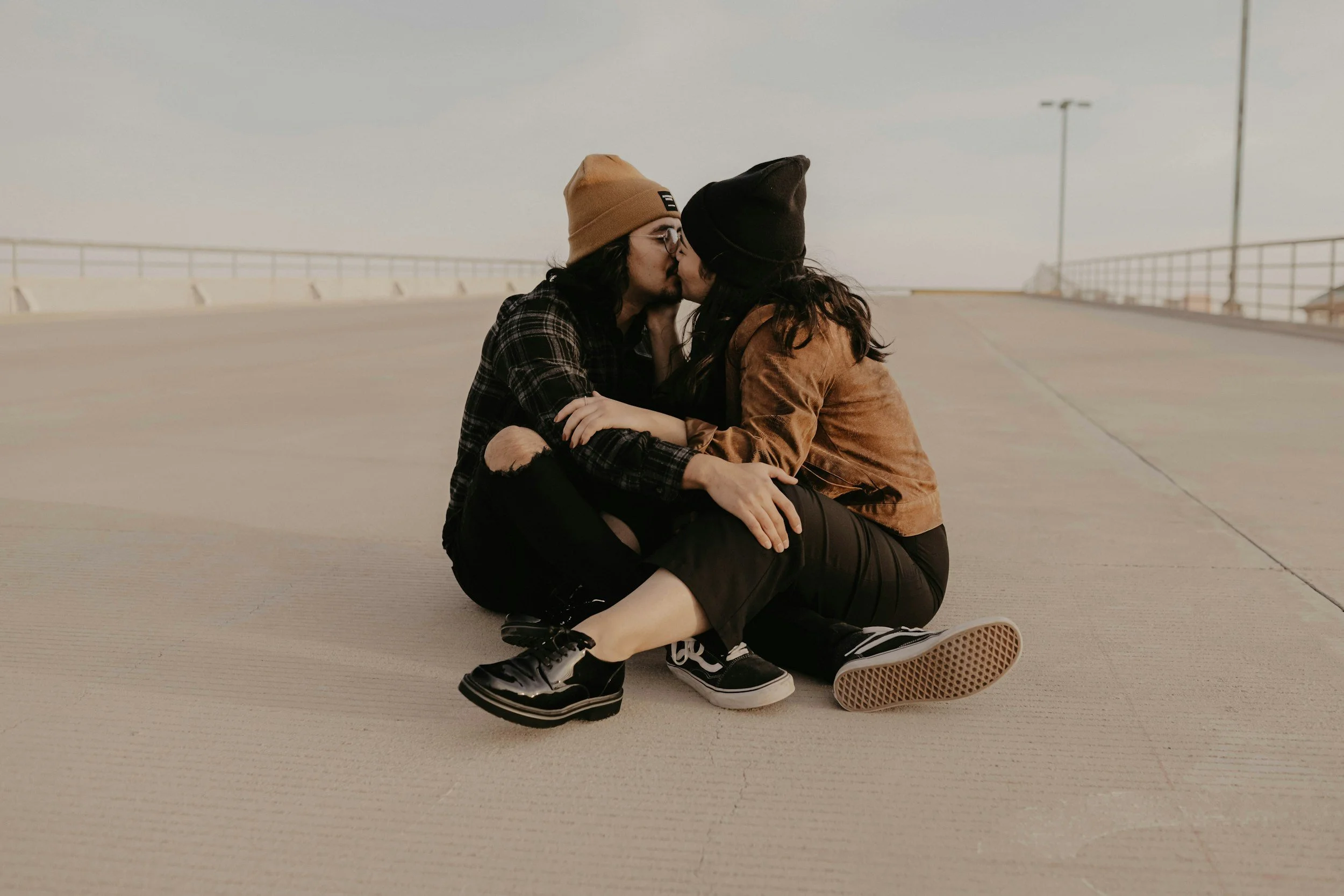 Two women sitting on a concrete surface sharing a kiss, wearing casual clothing and beanies.
