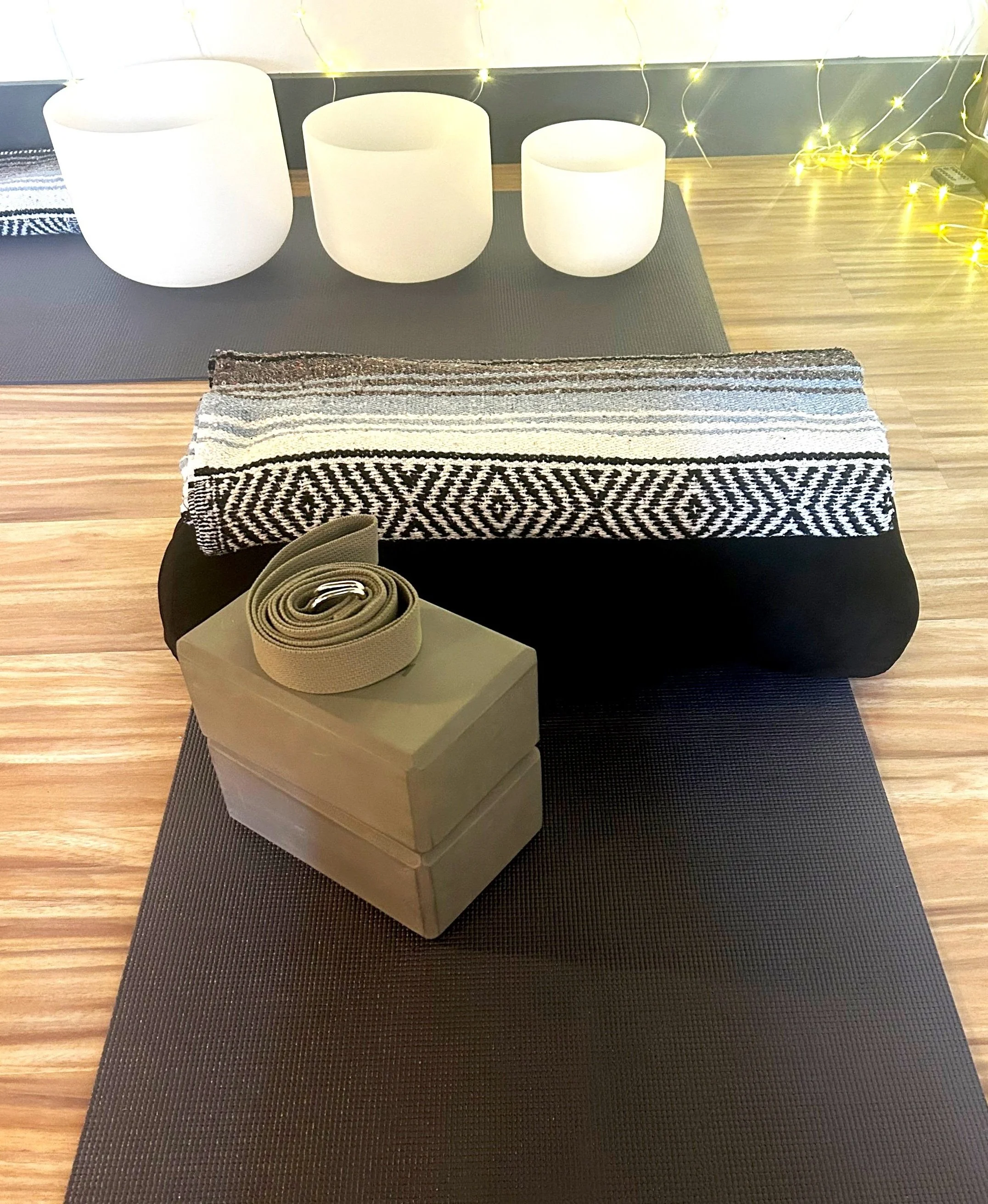 Yoga mats, a rolled blanket, and a foam block with a yoga belt on top, with white singing bowls and string lights in the background.