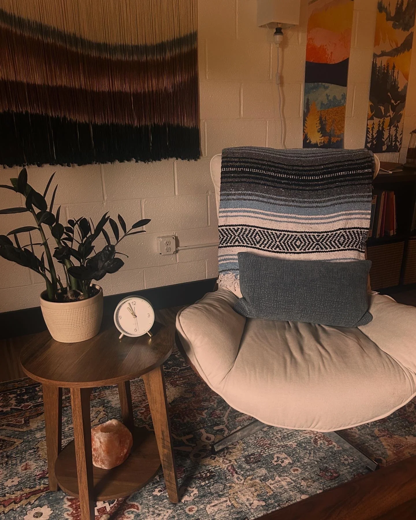 A cozy corner with a beige cushioned chair holding two pillows, a wooden side table with a plant, a clock, and a salt rock. There's a textured wall hanging, abstract wall art, and a bookshelf in the background.