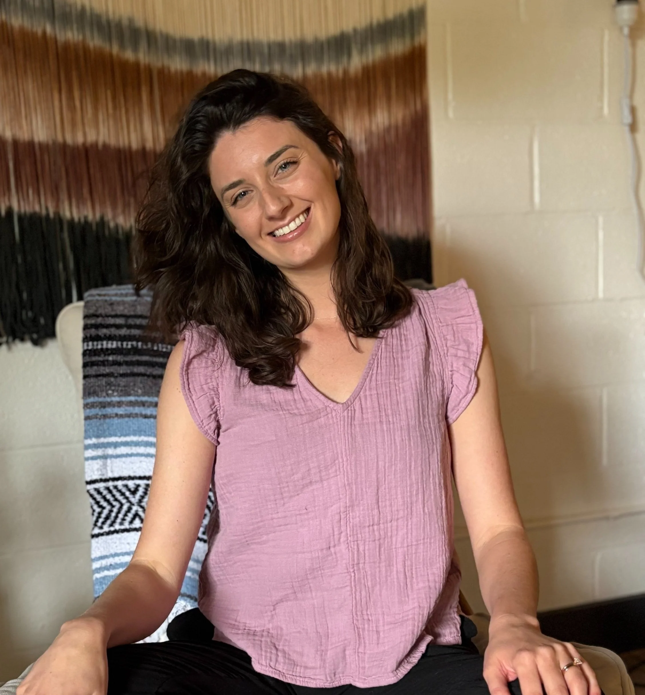 Smiling woman with shoulder-length curly brown hair wearing a pink sleeveless top, sitting indoors on a chair with a black and white woven blanket in the background.