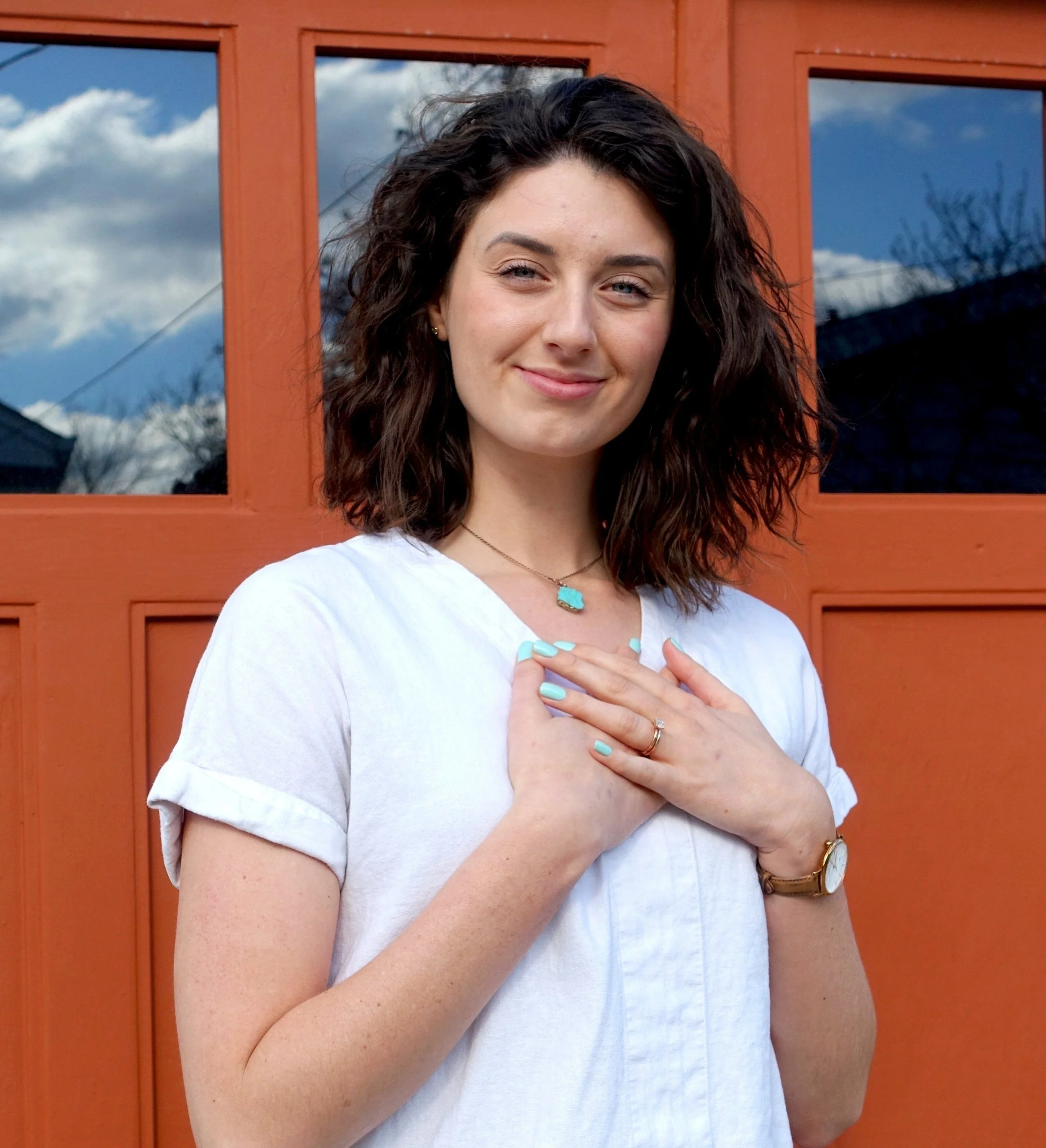 A young woman with curly brown hair, wearing a white shirt, a turquoise necklace, and a watch, standing in front of a red door with windows reflecting blue skies and clouds.