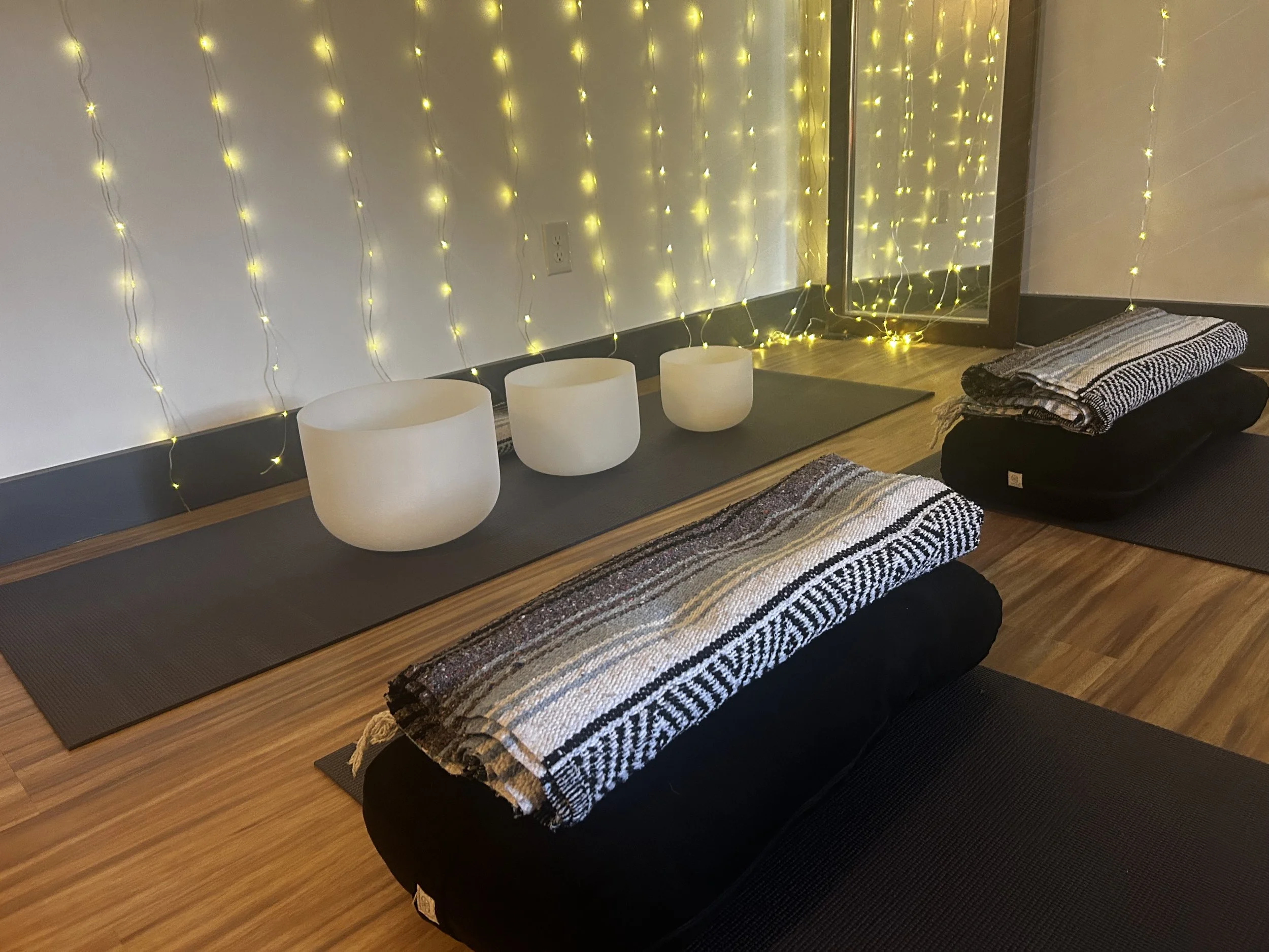 Yoga mats with folded blankets, white singing bowls, and yellow string lights on a wall reflect off a glass door.