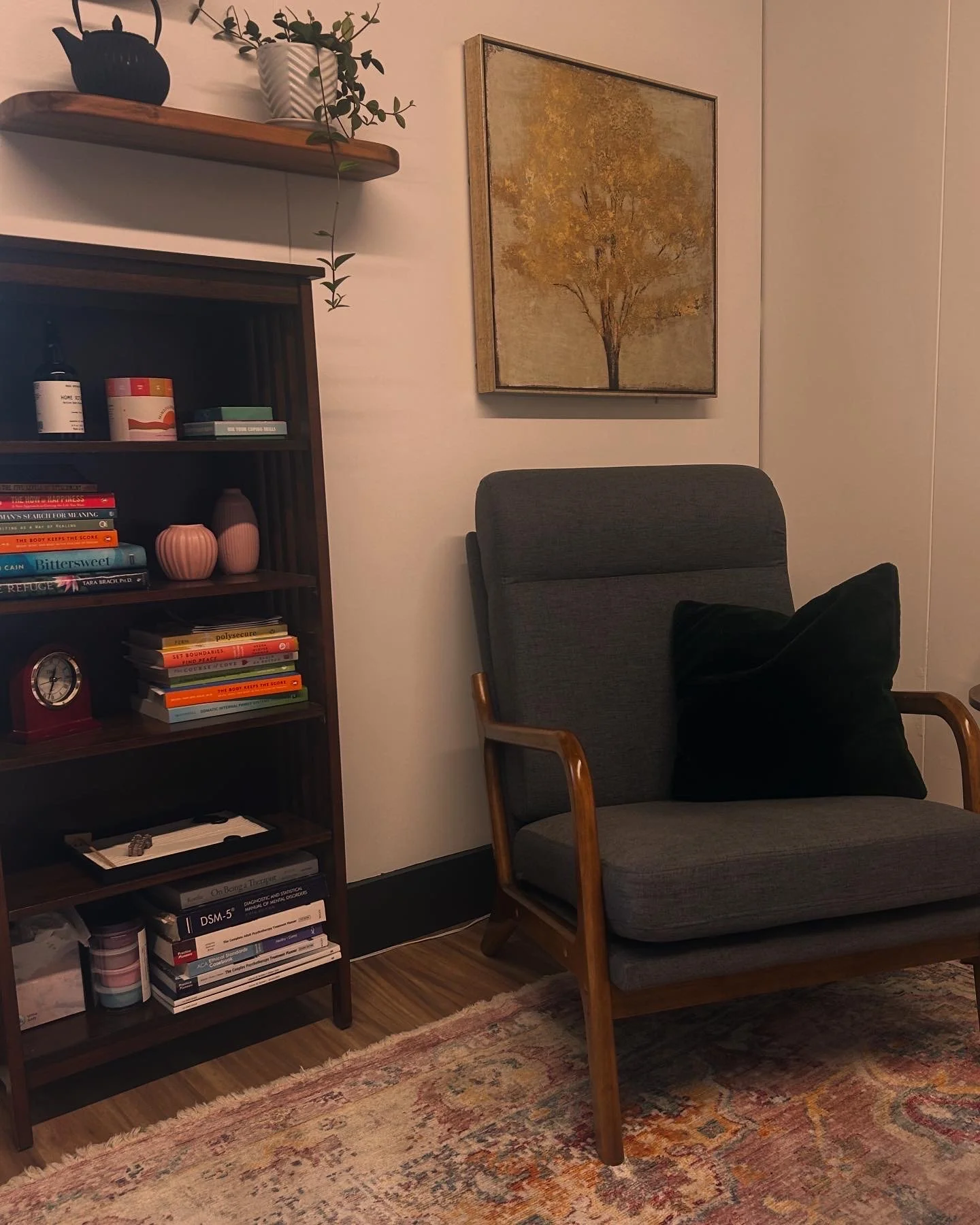 A cozy corner of a living room with a gray armchair, black pillow, wooden armrests, a bookshelf filled with books and decor, a wall painting of a tree, and a wooden shelf with potted plants above the bookshelf.