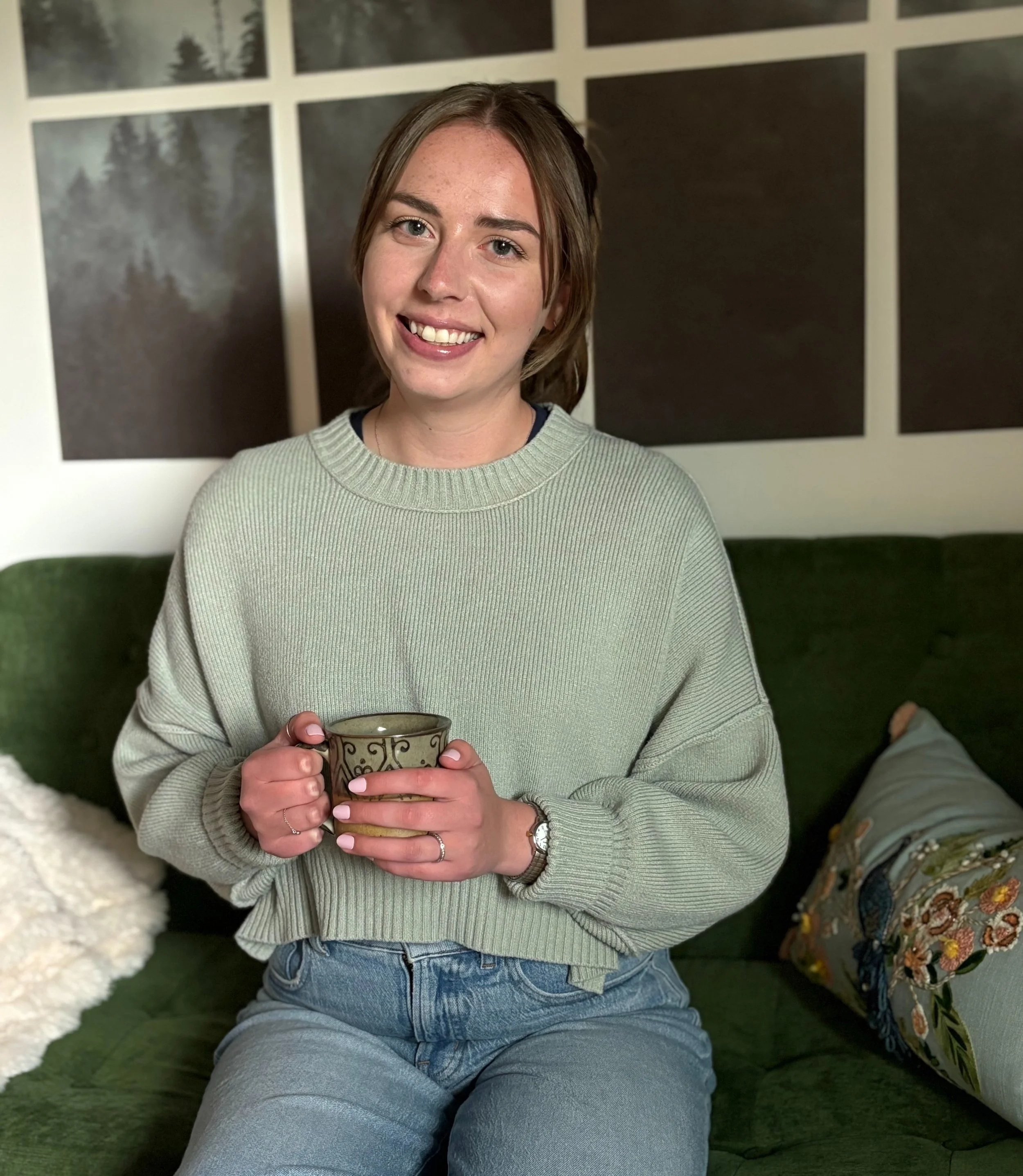A young woman with light brown hair, wearing a light green sweater and blue jeans, sitting on a green couch, holding a patterned mug, smiling at the camera. In the background, framed pictures of trees are visible on the wall, and there are decorative pillows on the couch.