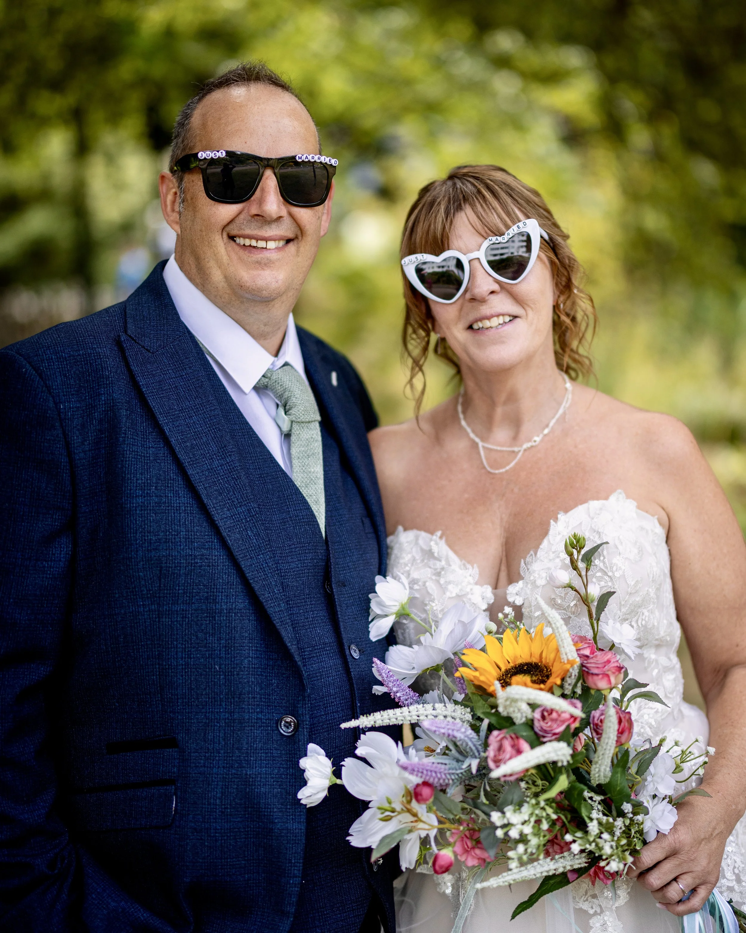 A smiling bride and groom wearing sunglasses, with the bride holding a colorful bouquet, outdoors in a green, leafy setting.