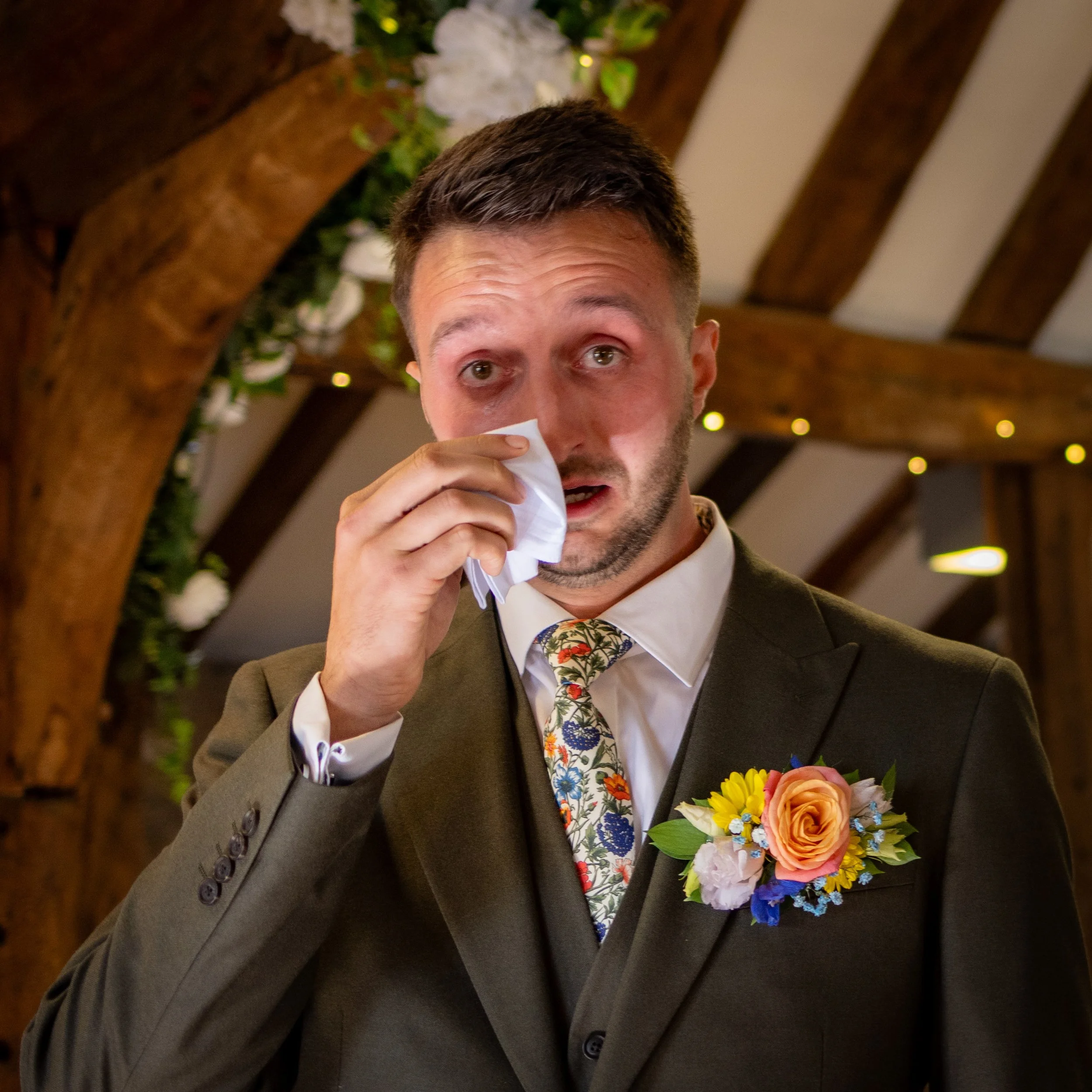 A man in a suit with a floral tie and a boutonniere is emotional, holding a tissue to his nose, possibly tearing up, at an indoor event with wooden beams and fairy lights.