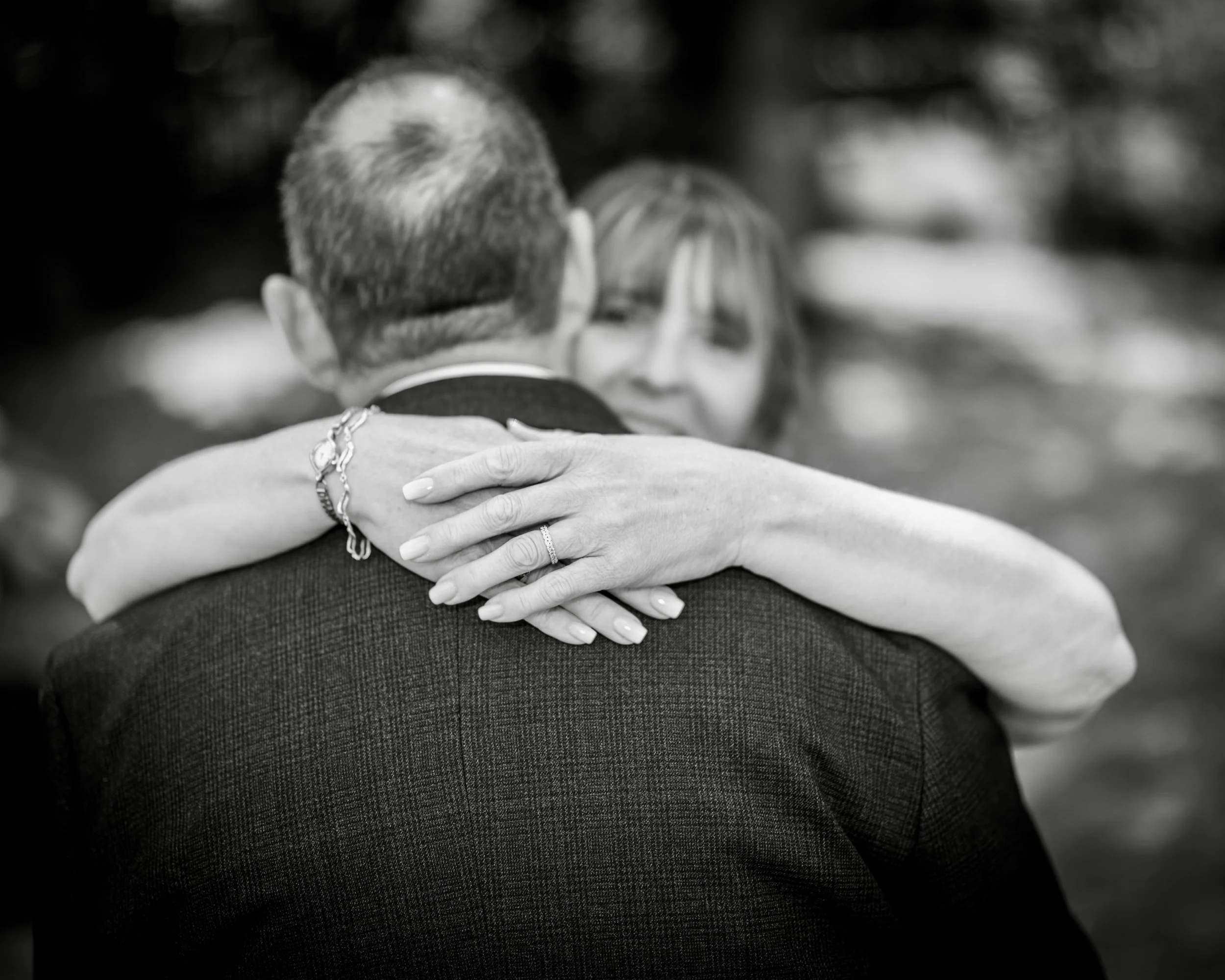 A person with short hair wearing a suit embraces another person, their arms around each other's shoulders in a hug.