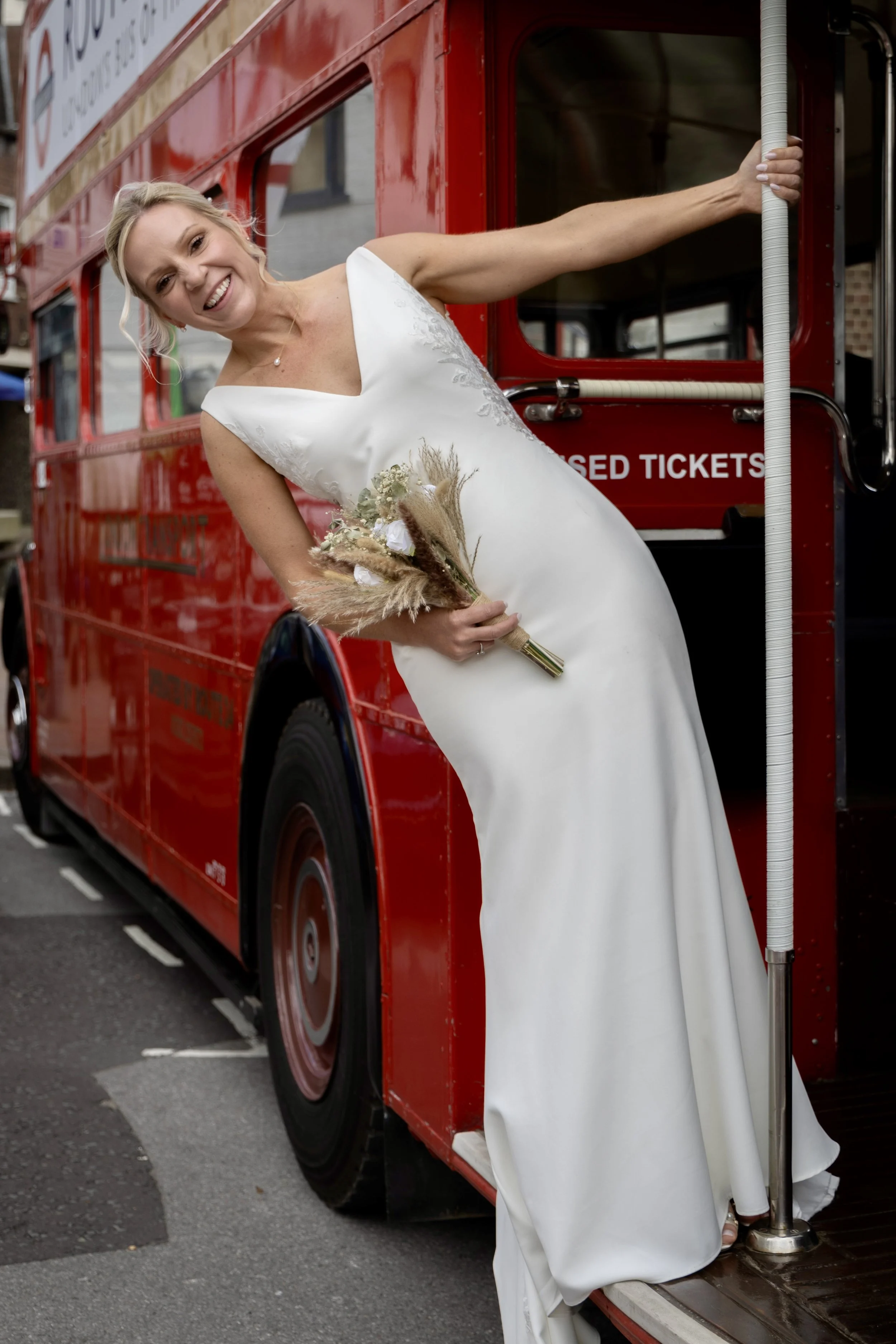 A woman in a white dress is smiling and stepping onto a red double-decker bus, holding a bouquet of flowers.