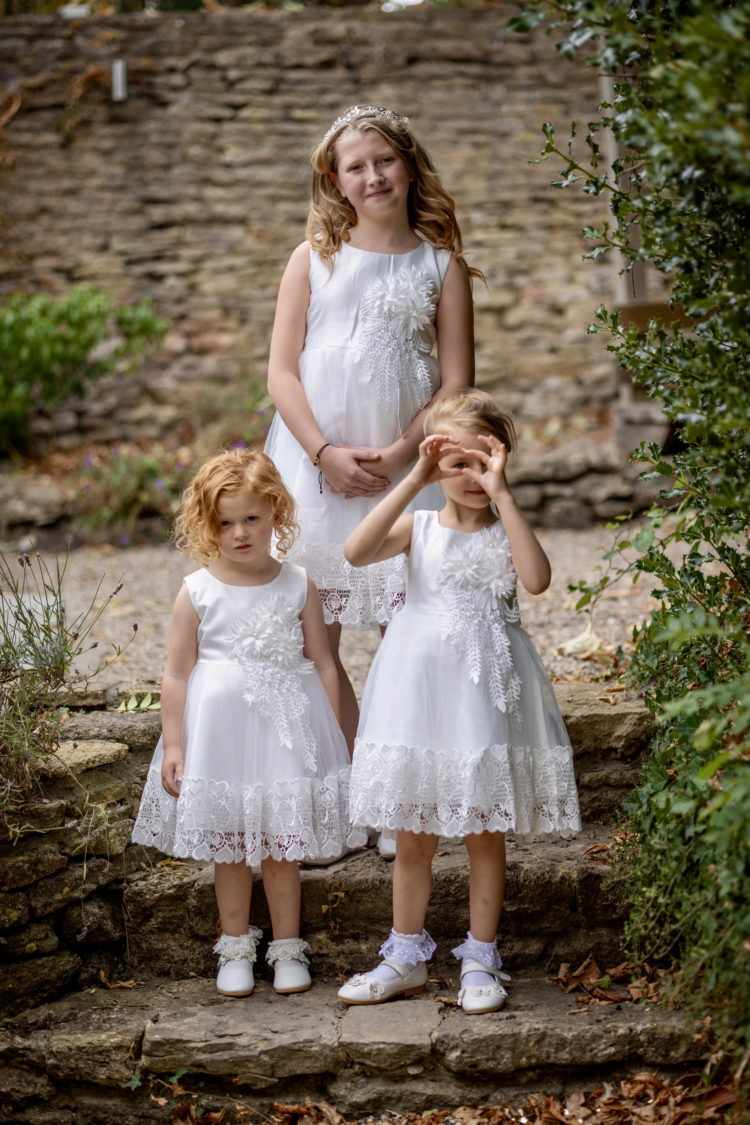 Four young girls dressed in white lace dresses, standing outdoors on stone stairs with green foliage around, posing for a photo.