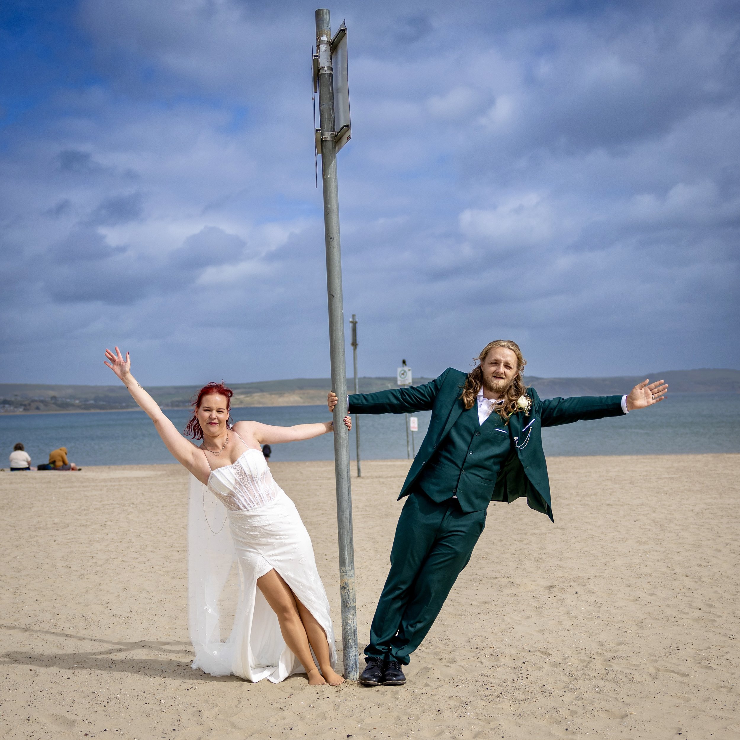 A bride and groom are standing on the beach with arms outstretched, holding a pole with a blank sign, under a cloudy sky. Part of the bride's white wedding dress is visible, and the groom is in a green suit.