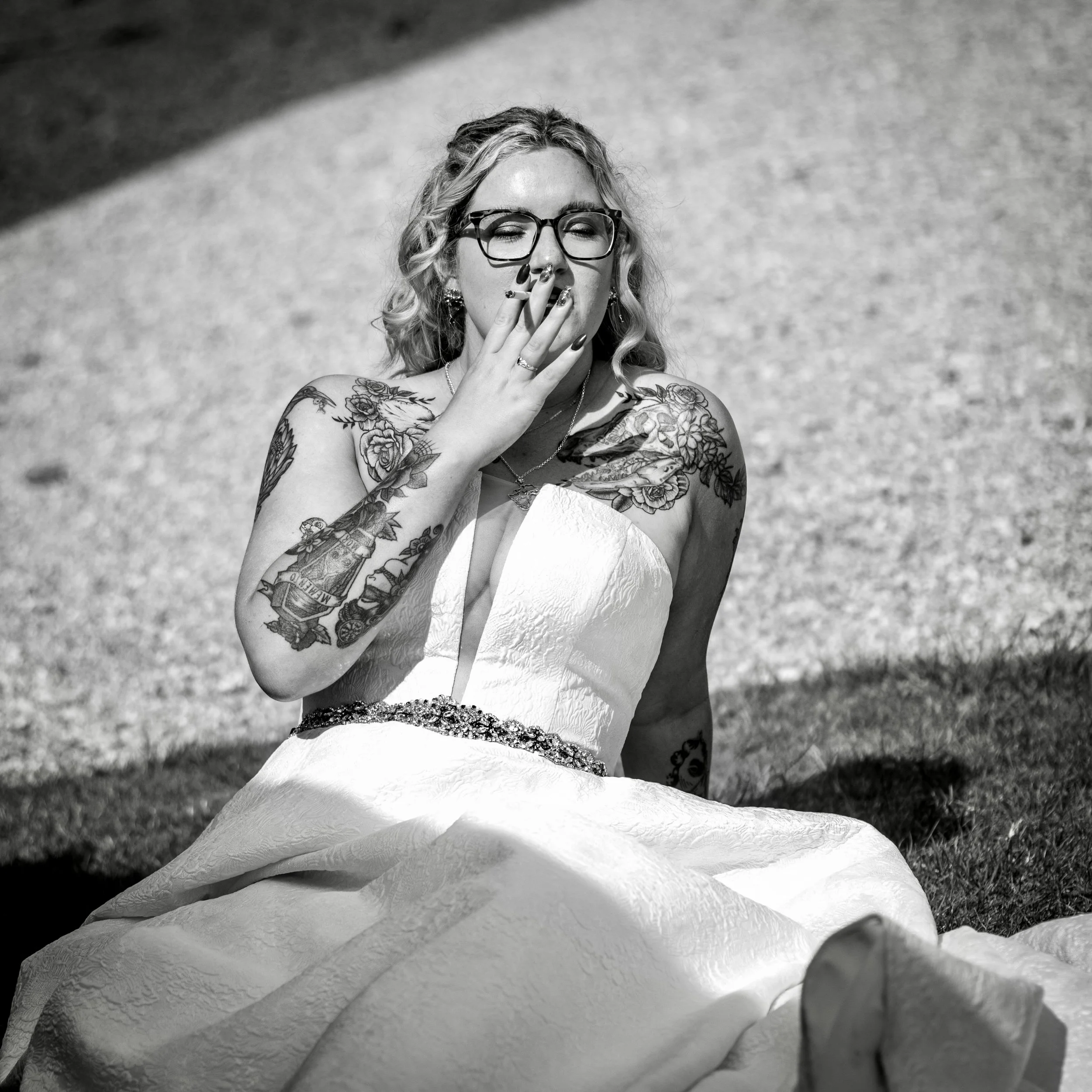 Black and white photo of a woman with glasses, tattoos on her arm, and jewelry, holding a cigarette to her mouth on a textured surface.