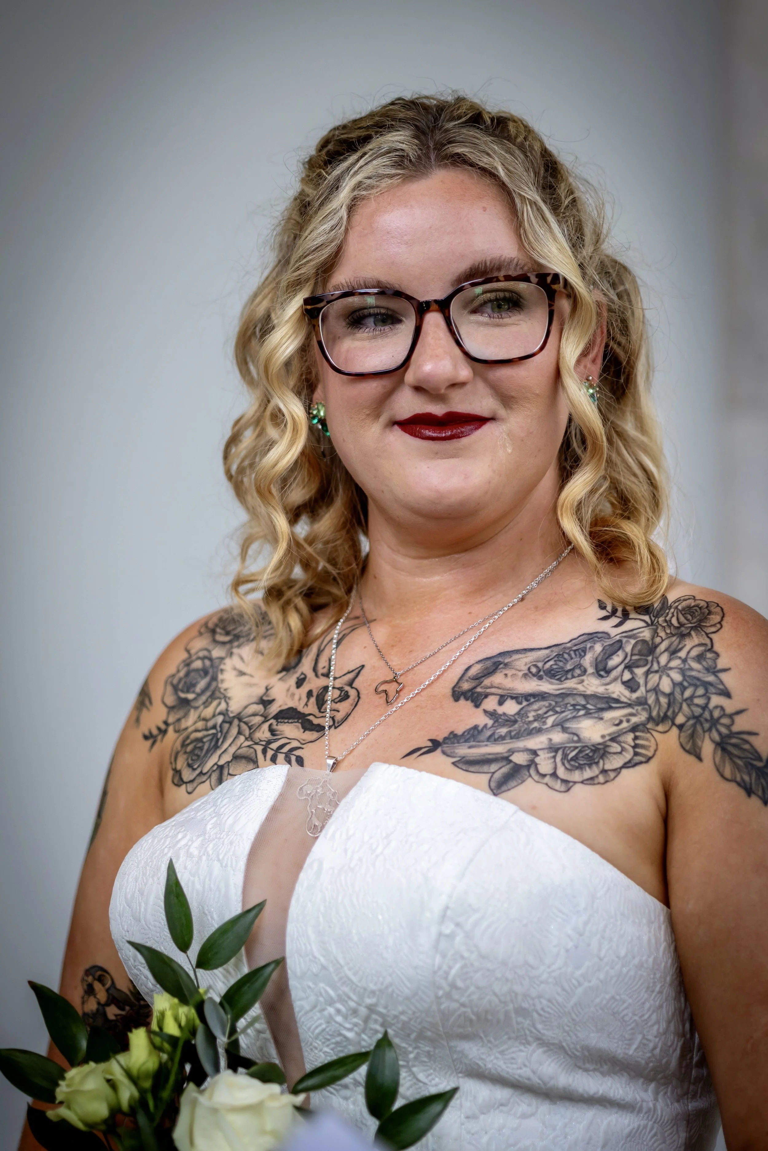 A woman with blonde curly hair wearing glasses, red lipstick, and earrings, holding a bouquet of white roses and greenery, dressed in a strapless white wedding dress with tattooed shoulders and chest, standing against a plain background.