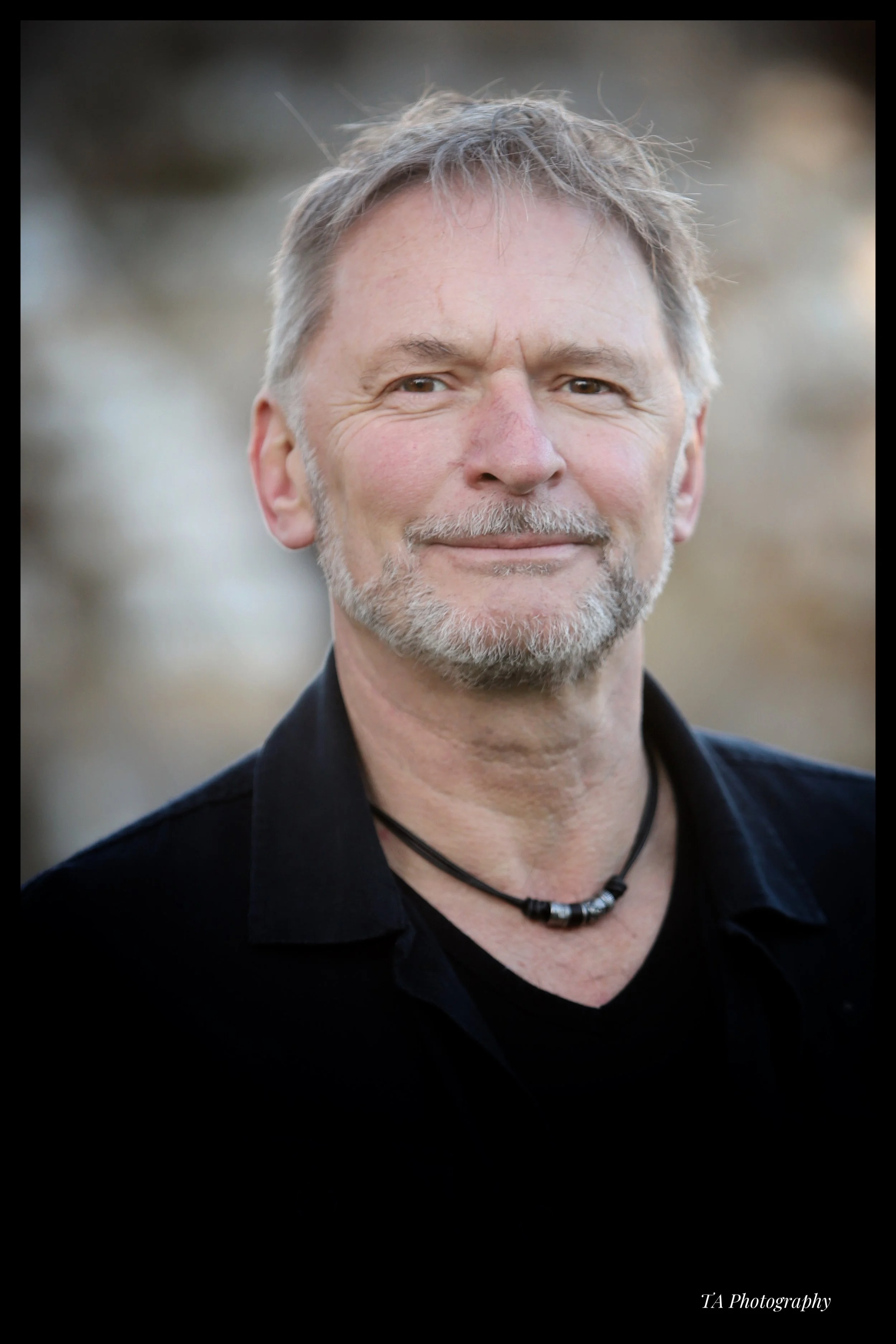 Close-up of a middle-aged man with short gray hair, beard, and light skin, smiling slightly, wearing a black shirt and a beaded necklace, outdoors with blurred background.