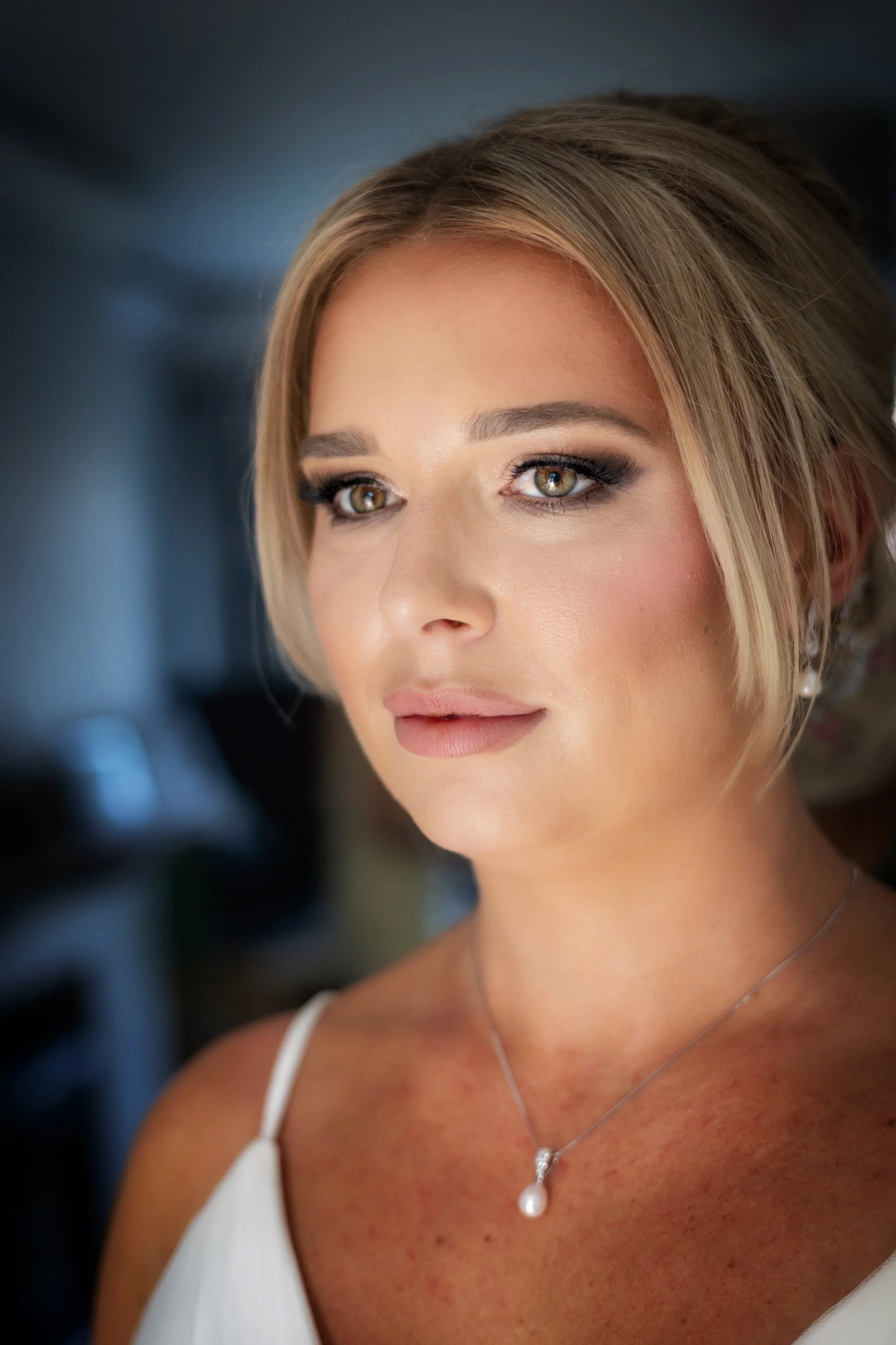 Close-up of a woman with blonde hair and hazel eyes, wearing makeup, a pearl necklace, and a white top, with a soft focus background.