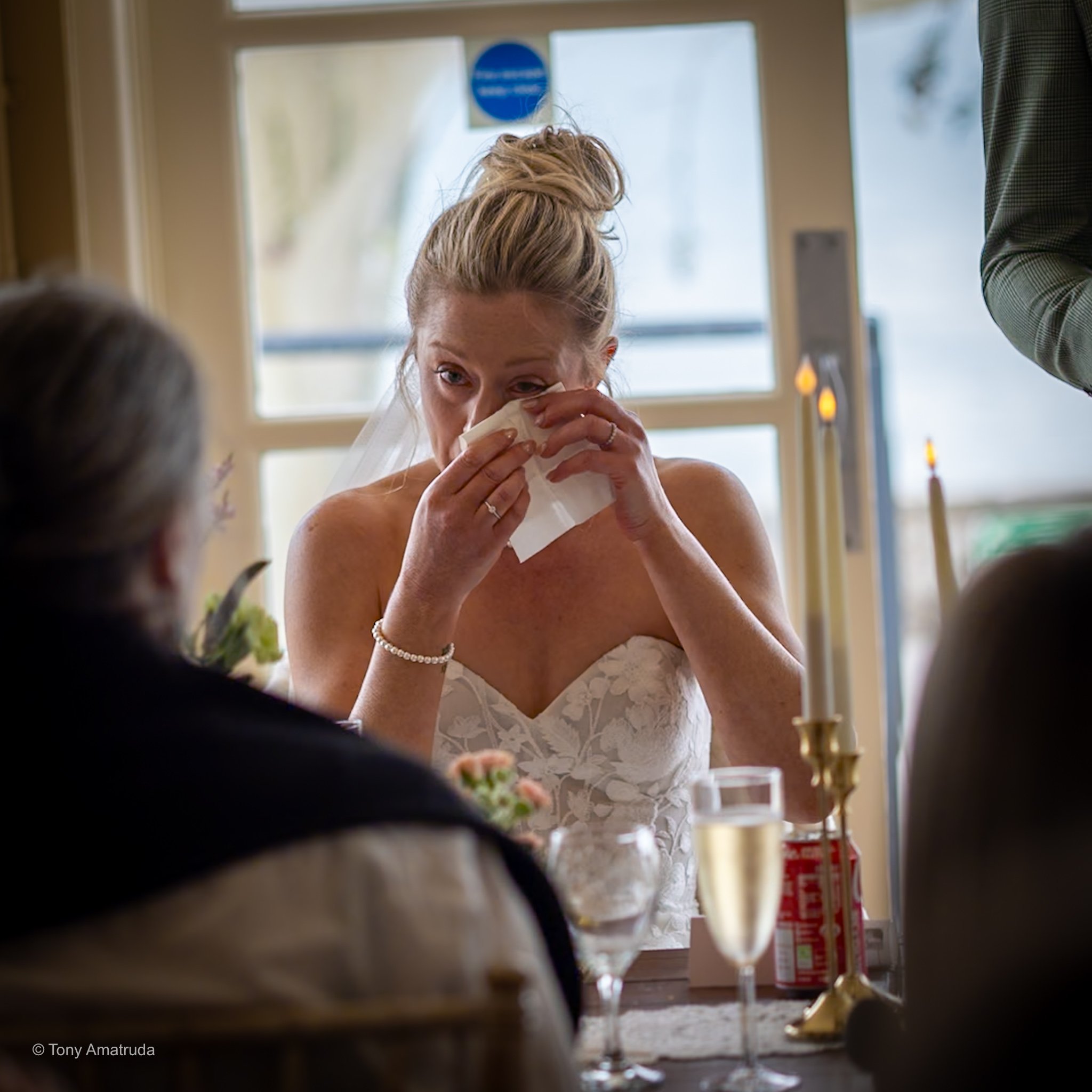 A woman with blonde hair tied in a bun, wearing a white strapless dress, is wiping her eye with a tissue inside a room with a window in the background. The scene appears to be from a wedding or special event.