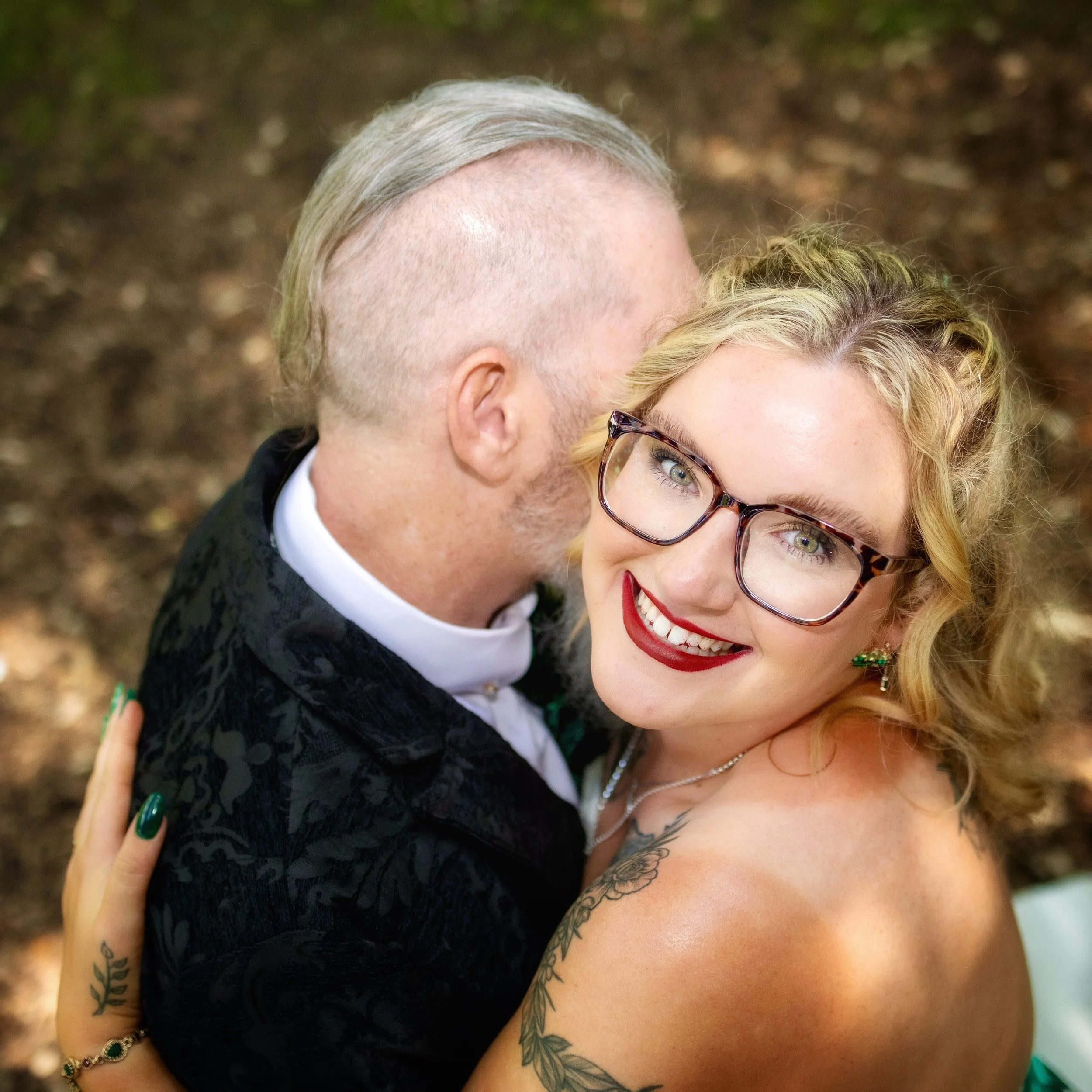 A smiling woman with curly blonde hair, glasses, and tattoos hugging a man with a gray beard, who has his face close to her cheek, outdoors with a blurred natural background.