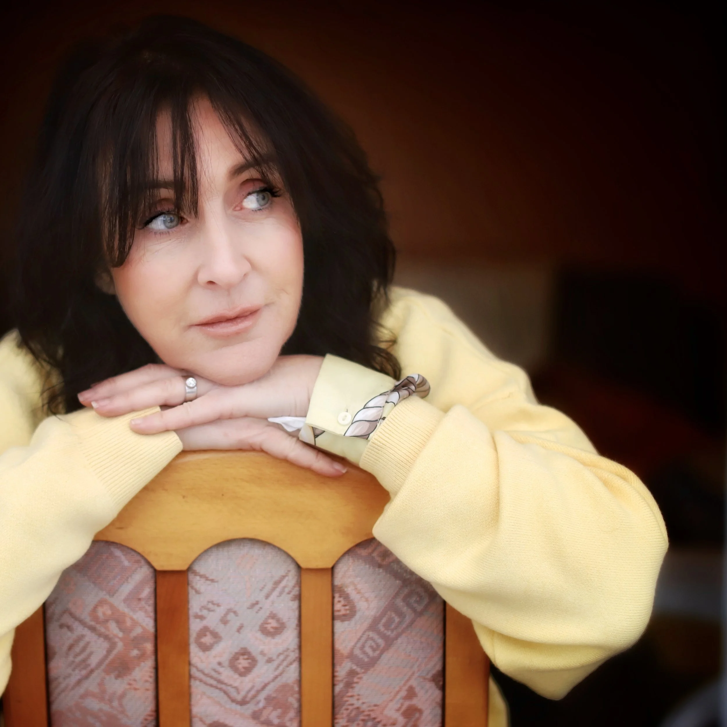 Close-up of a woman with shoulder-length dark hair, blue eyes, wearing a yellow sweater, resting her chin on her hand, sitting at a wooden table with a tiled floor underneath.