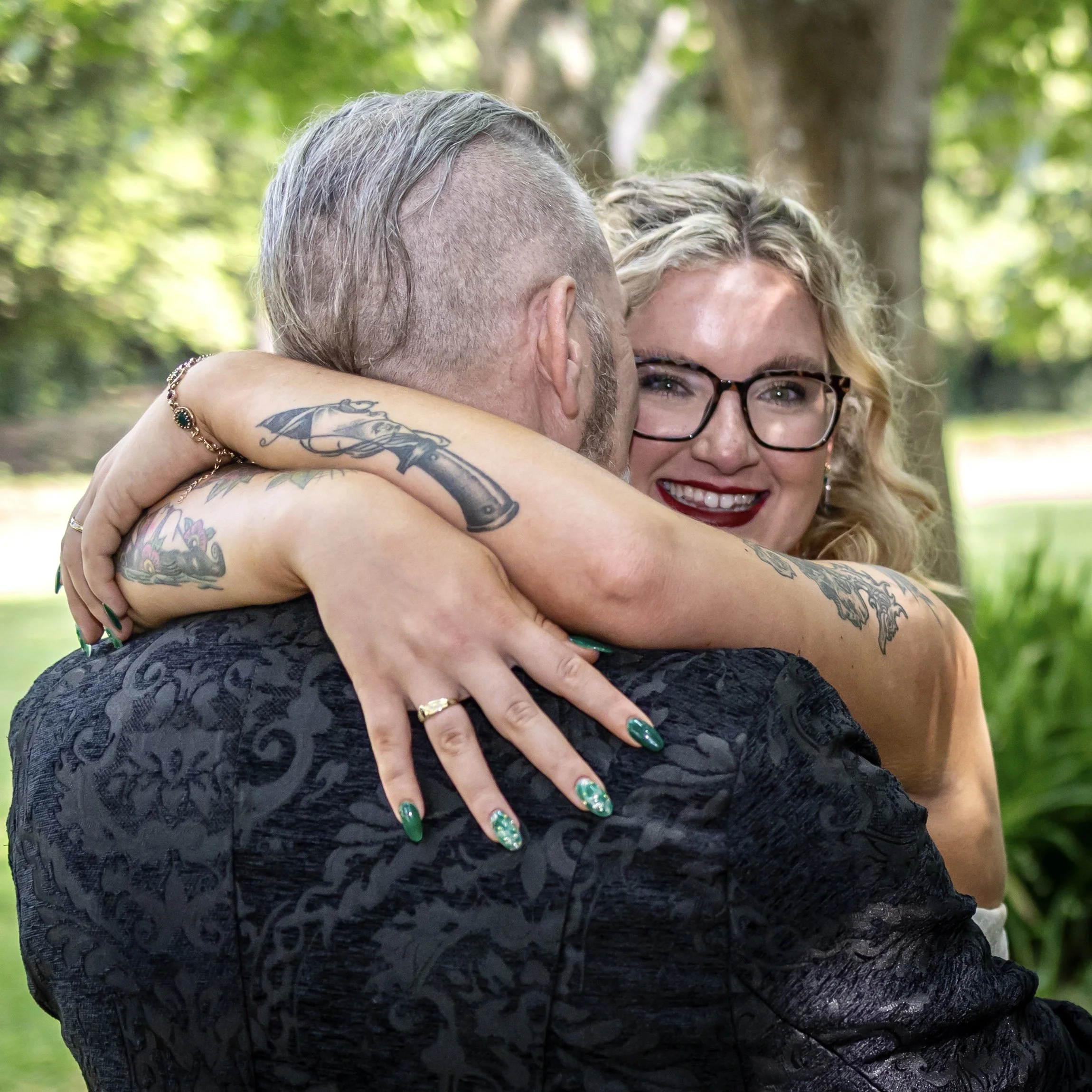 Close-up of a couple hugging outdoors, the woman wearing glasses, smiling, with tattoos on her arms, and the man with a hairstyle shaved on the sides and longer on top, in a green park with trees in the background.