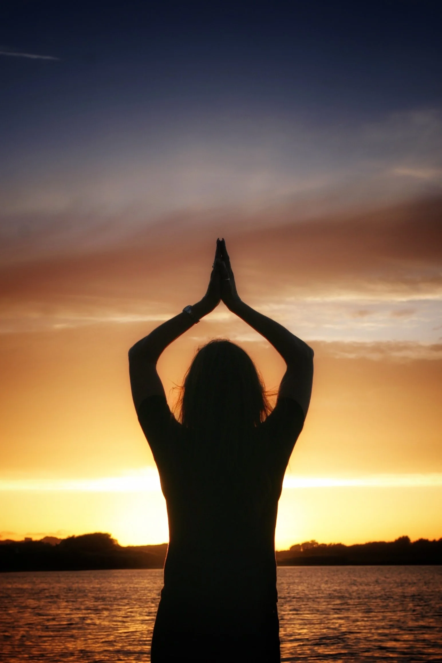 A person standing by a lake at sunset, silhouetted, with their hands in a prayer position above their head, facing the sunset.