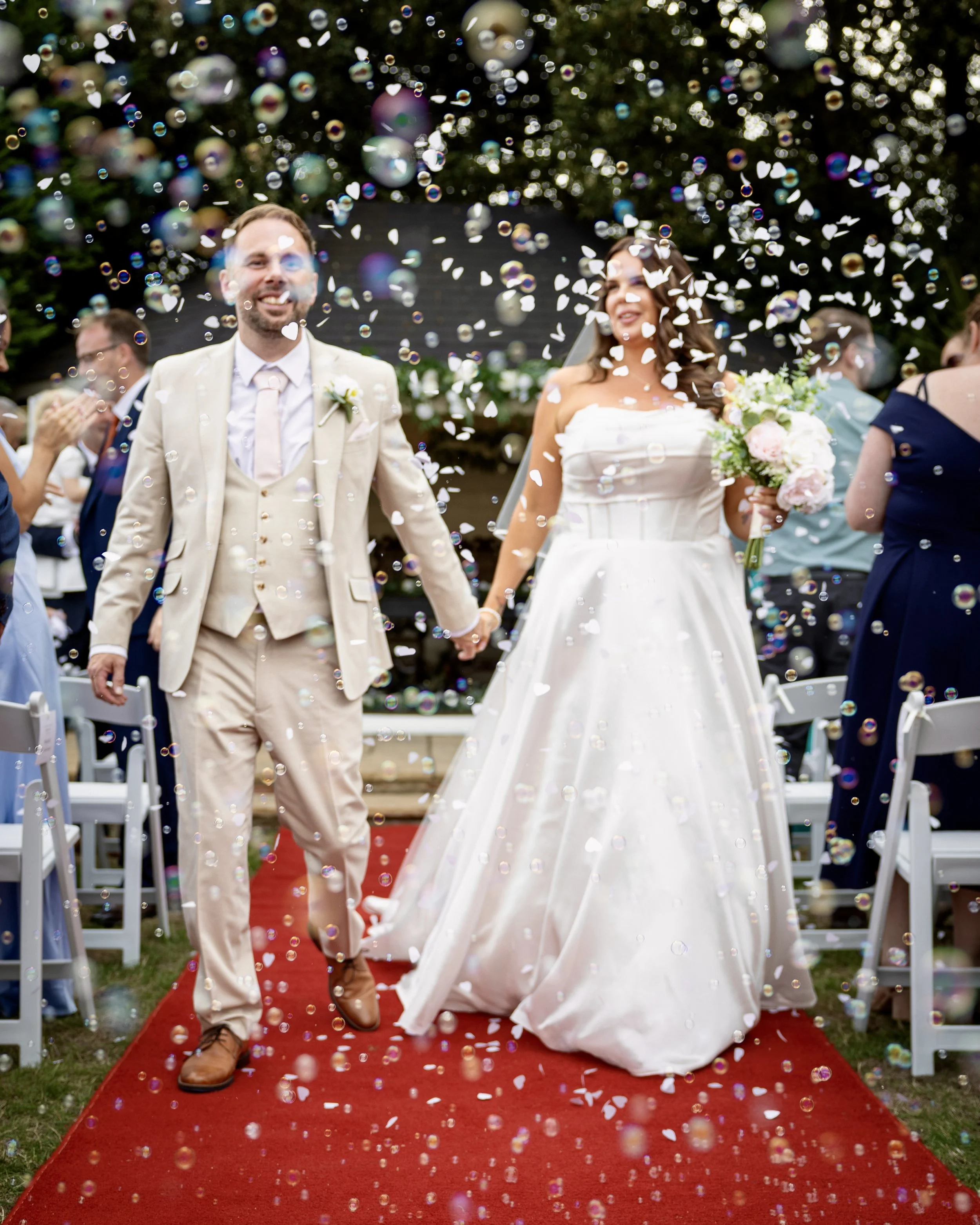 A newlywed couple holding hands and walking on a red carpet under falling confetti at an outdoor wedding celebration.