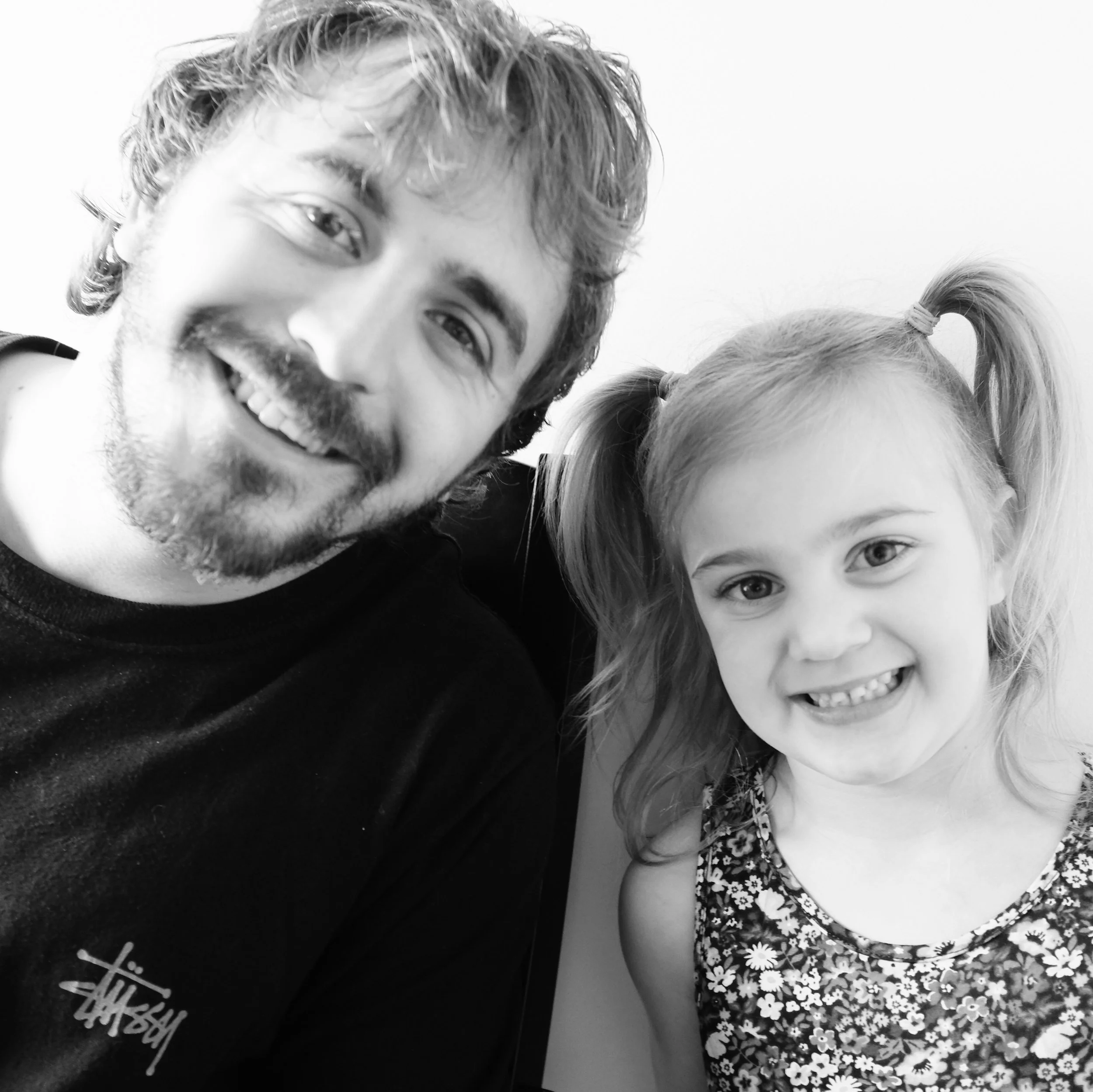 A man with curly hair and a beard smiling with a young girl with blonde hair in pigtails, wearing a floral dress, smiling widely.