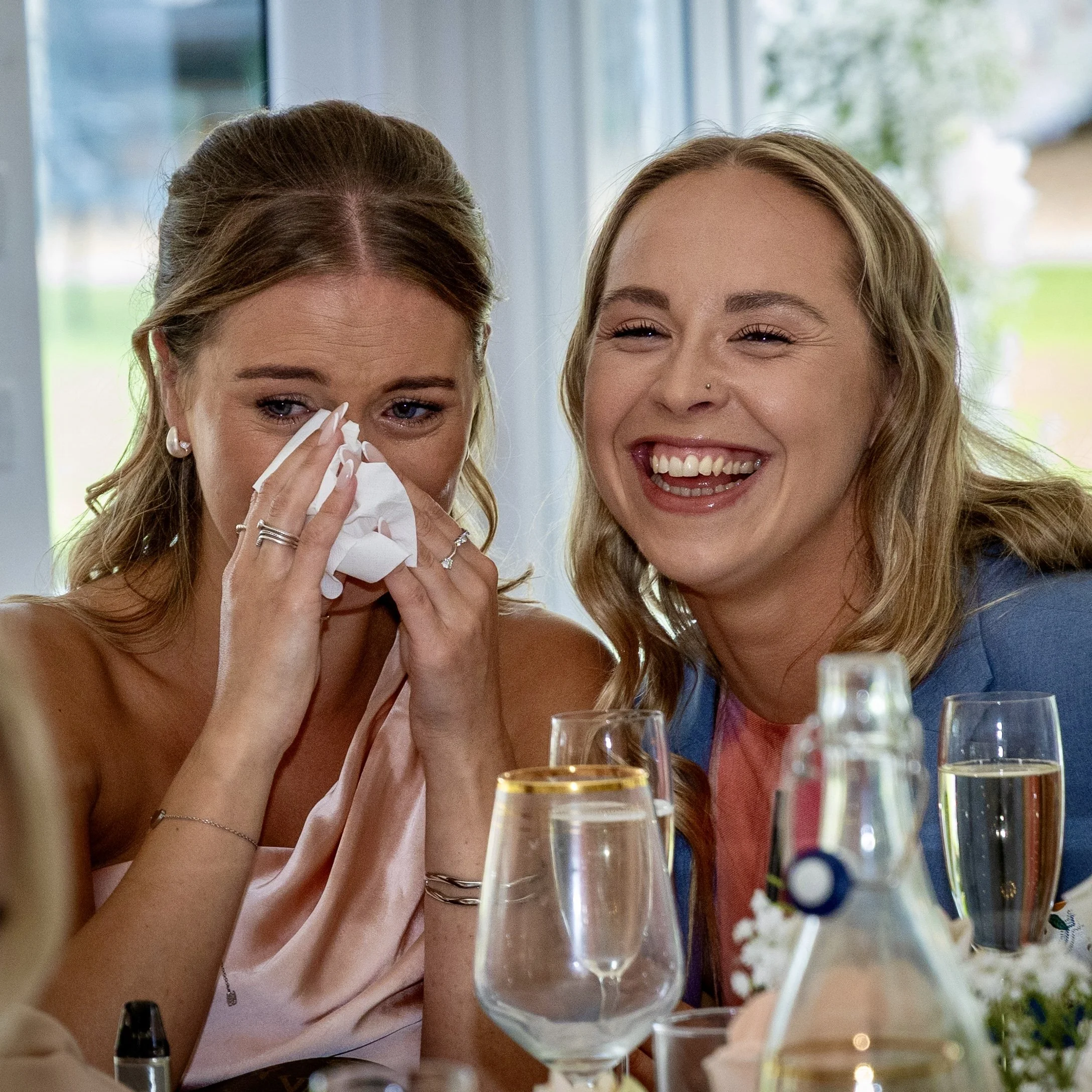 Two women sit at a table during a celebration, one is crying and wiping her eyes with a tissue, while the other is laughing and smiling. They are surrounded by glasses of drinks and a decorated table.