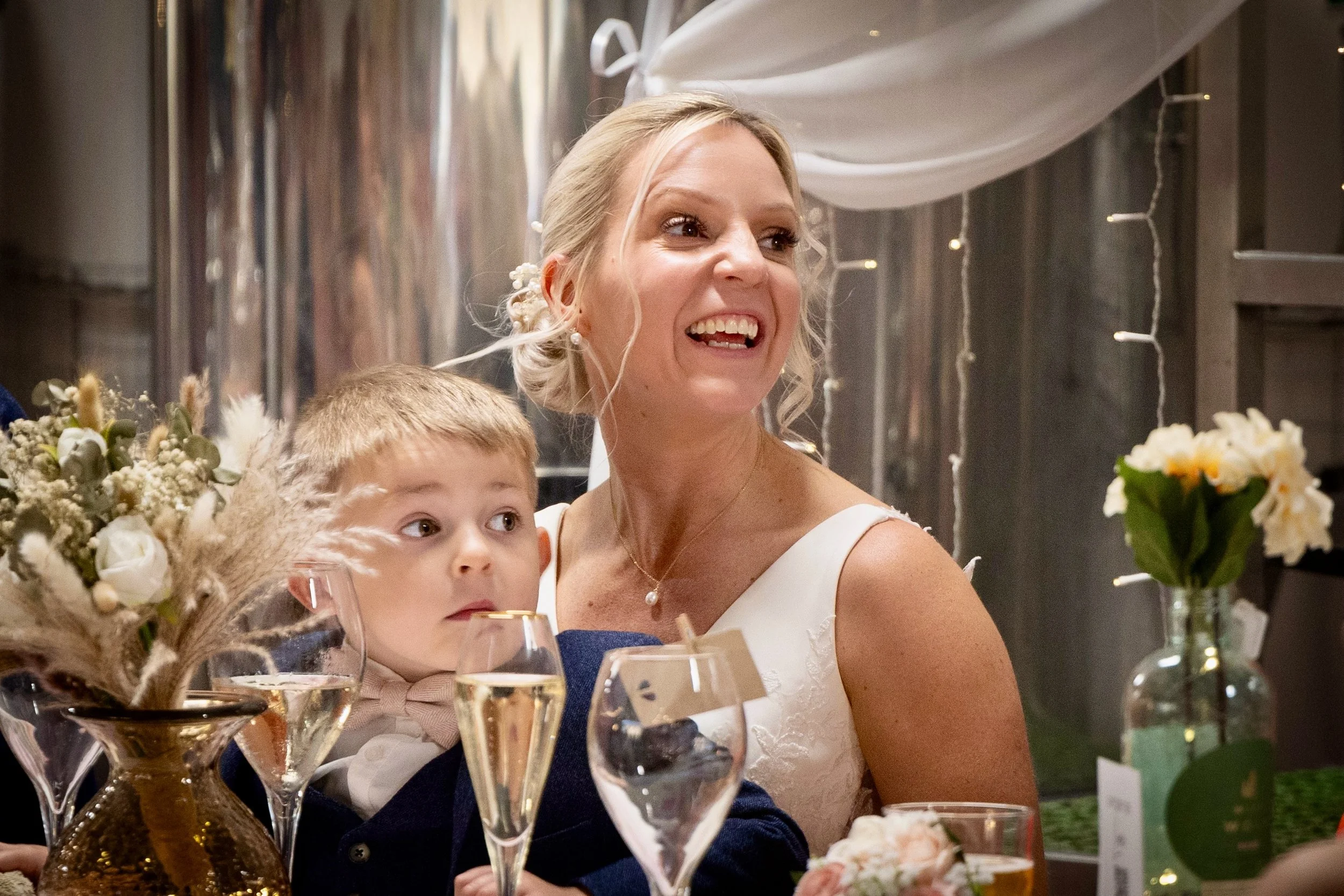 A woman with blonde hair smiling at a celebration or party, sitting next to a young boy at a table with flowers and drinks, decorated with string lights and white drapes in the background.