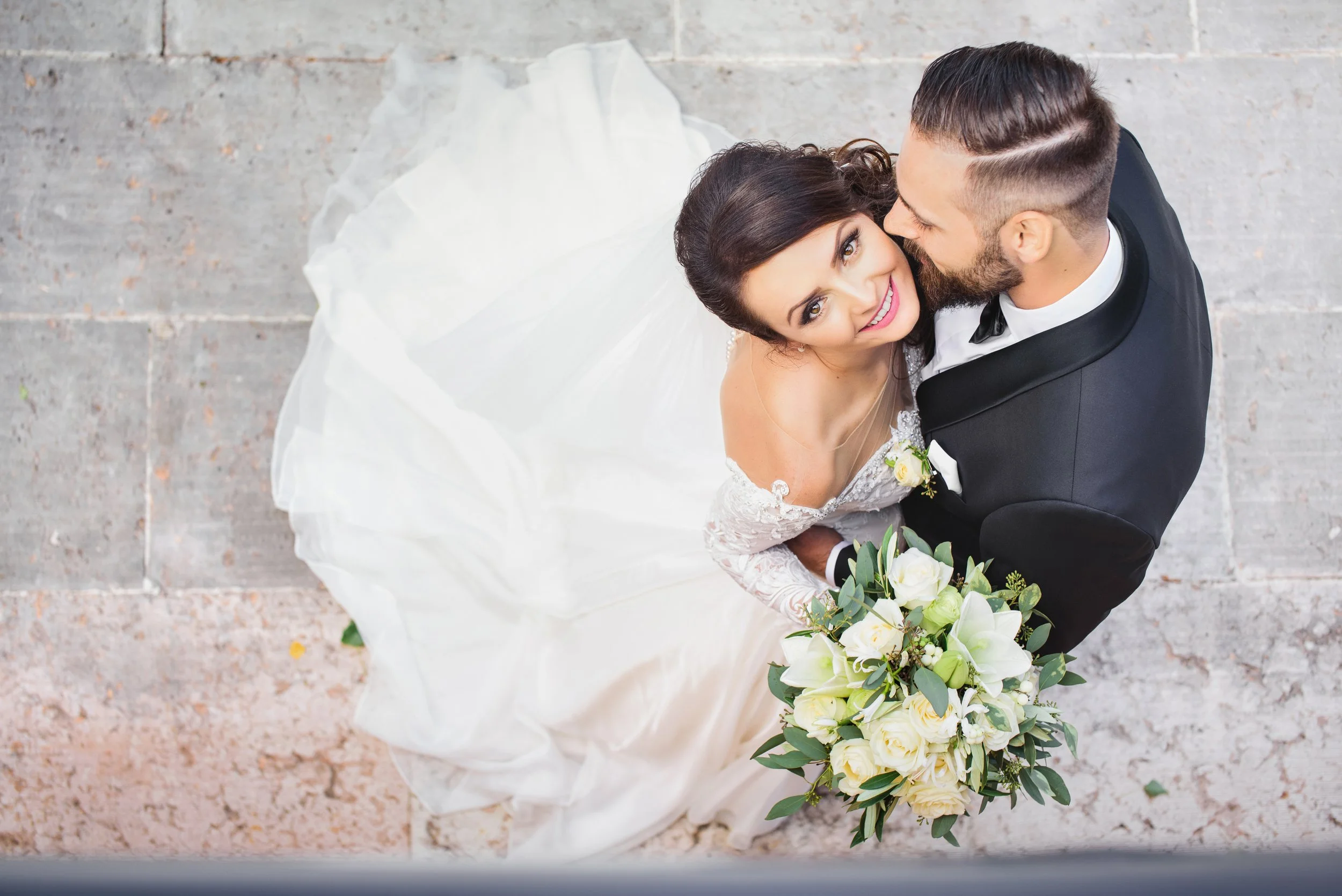 A bride and groom in wedding attire, looking up at the camera, with the bride holding a bouquet of white roses and greenery, standing on a tile floor.