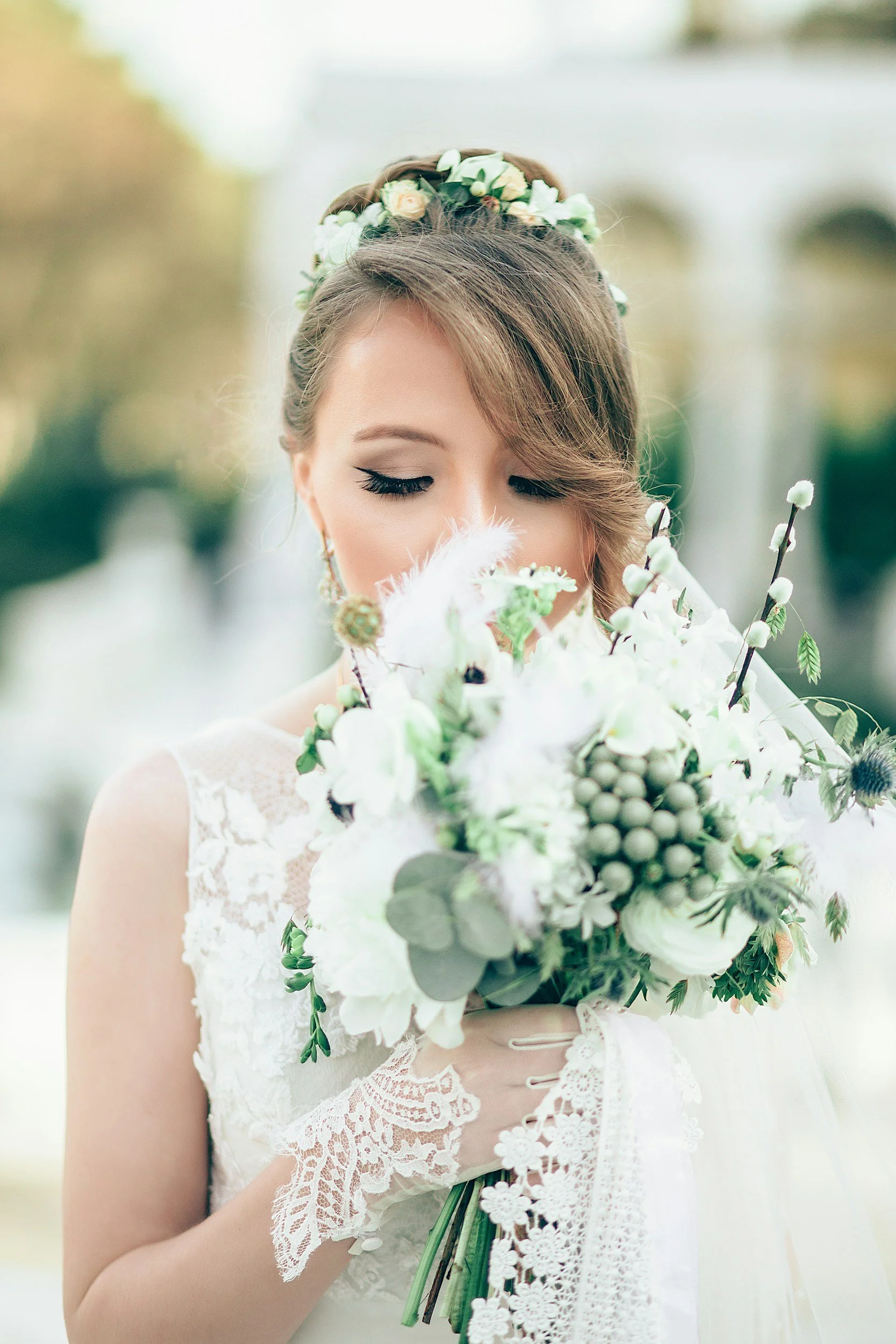 A bride with a floral crown holding a bouquet of white and green flowers, wearing lace gloves and a lace wedding dress, with her eyes closed.