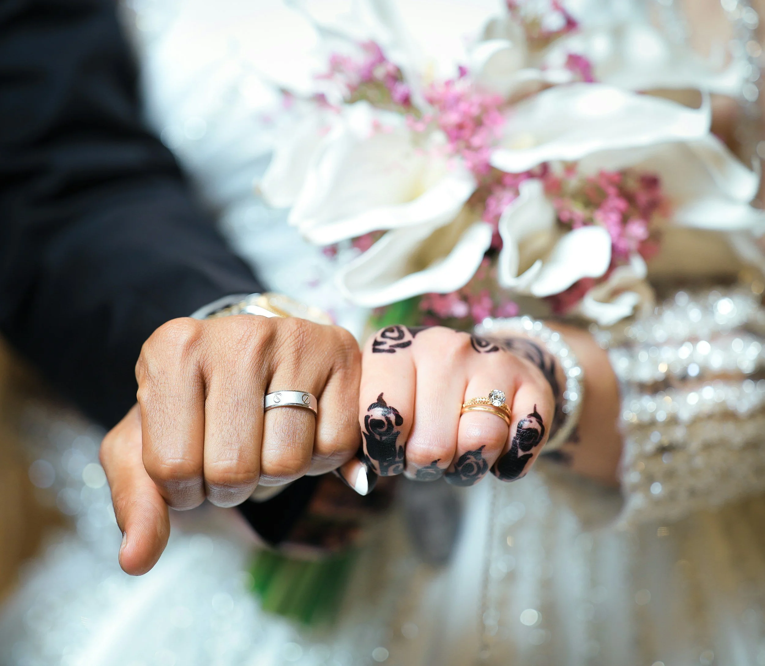Close-up of a person's hand showing multiple rings, with tattooed fingers, pointing downward against a background of white and pink flowers.