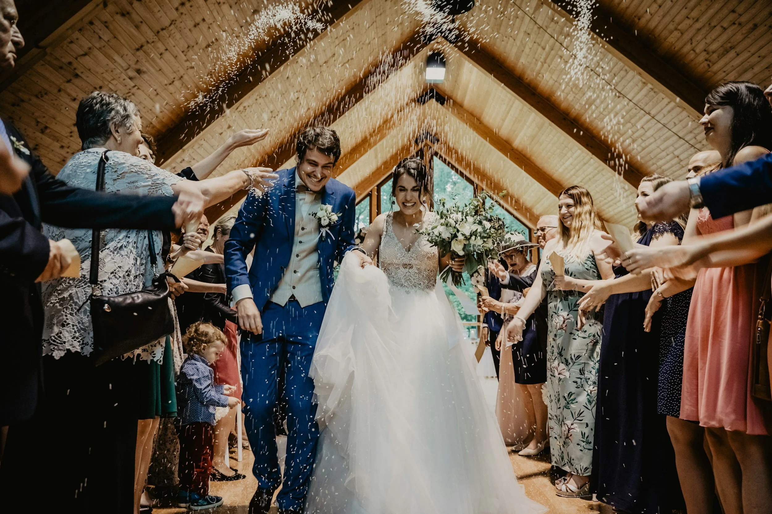A bride and groom walking together through a tunnel of friends and family throwing white rice during their wedding celebration inside a wooden venue.
