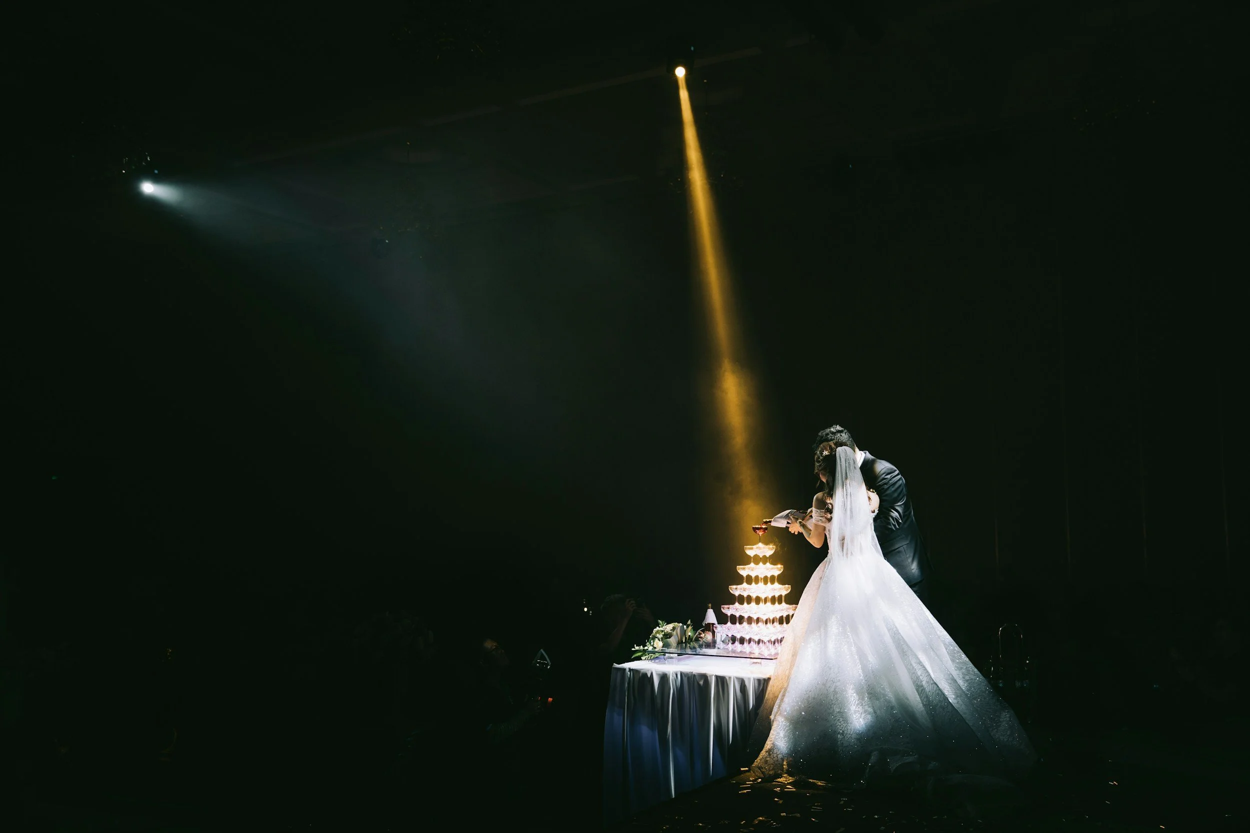 A couple dressed in wedding attire sharing a kiss during their wedding ceremony, with a tiered wedding cake on the table nearby, set against a dark background illuminated by a spotlight.