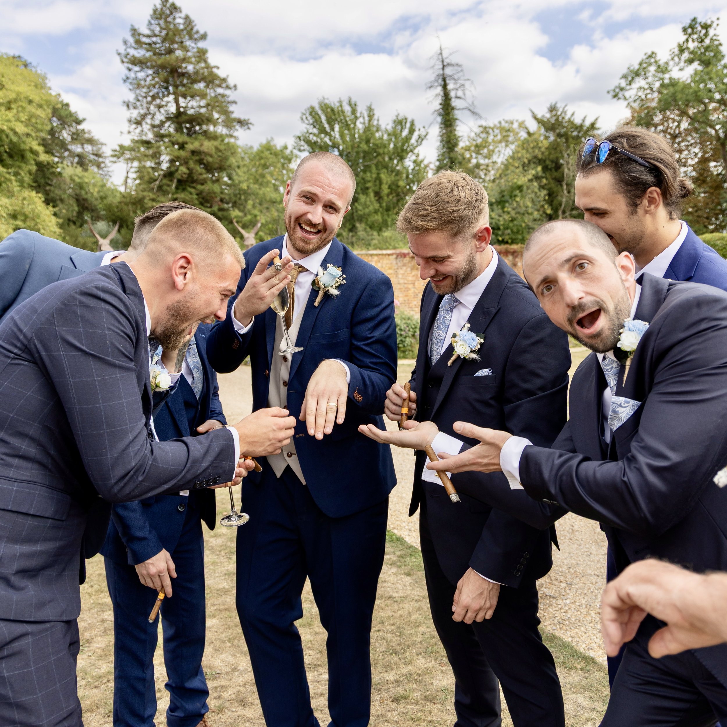 A group of six men dressed in suits, celebrating outdoors with drinks and cigars, smiling and laughing together on a cloudy day.