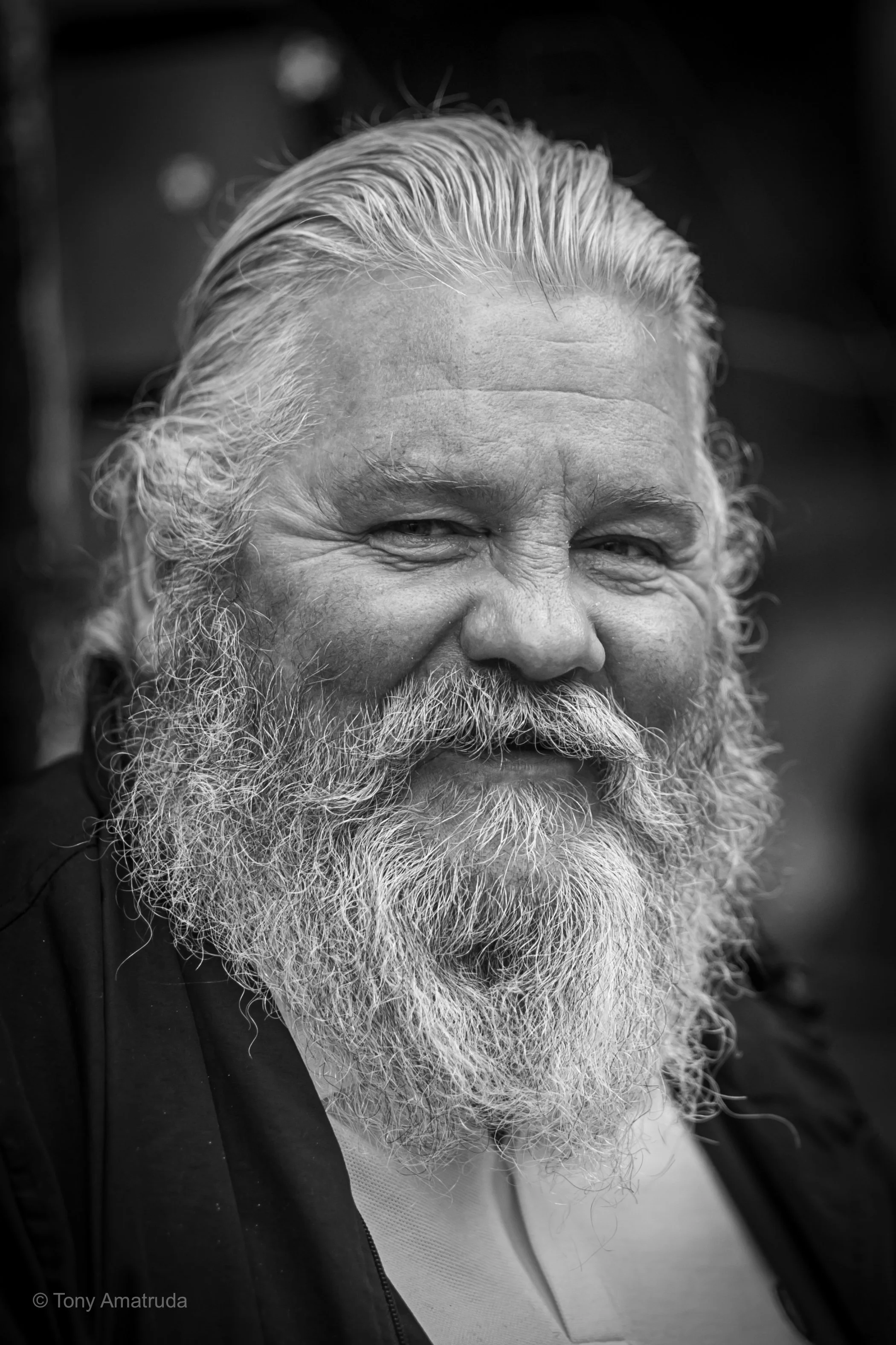 A close-up black and white photograph of an older man with a full white beard, mustache, and slicked-back hair, smiling slightly.
