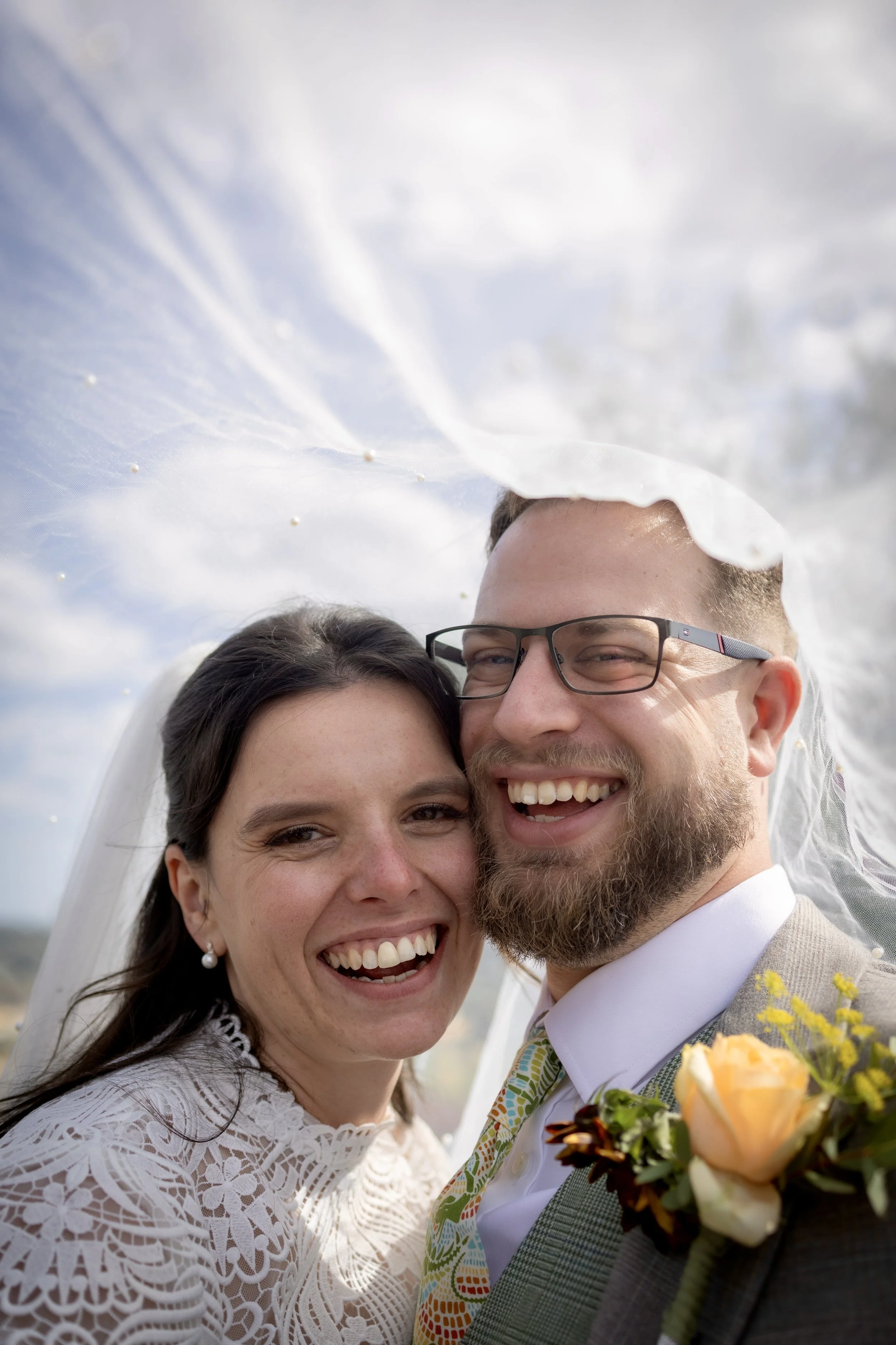 Close-up of a happy bride and groom smiling at their wedding, with the bride wearing a white lace dress and the groom in a checkered suit with a boutonniere and colorful tie, outdoors on a cloudy day.