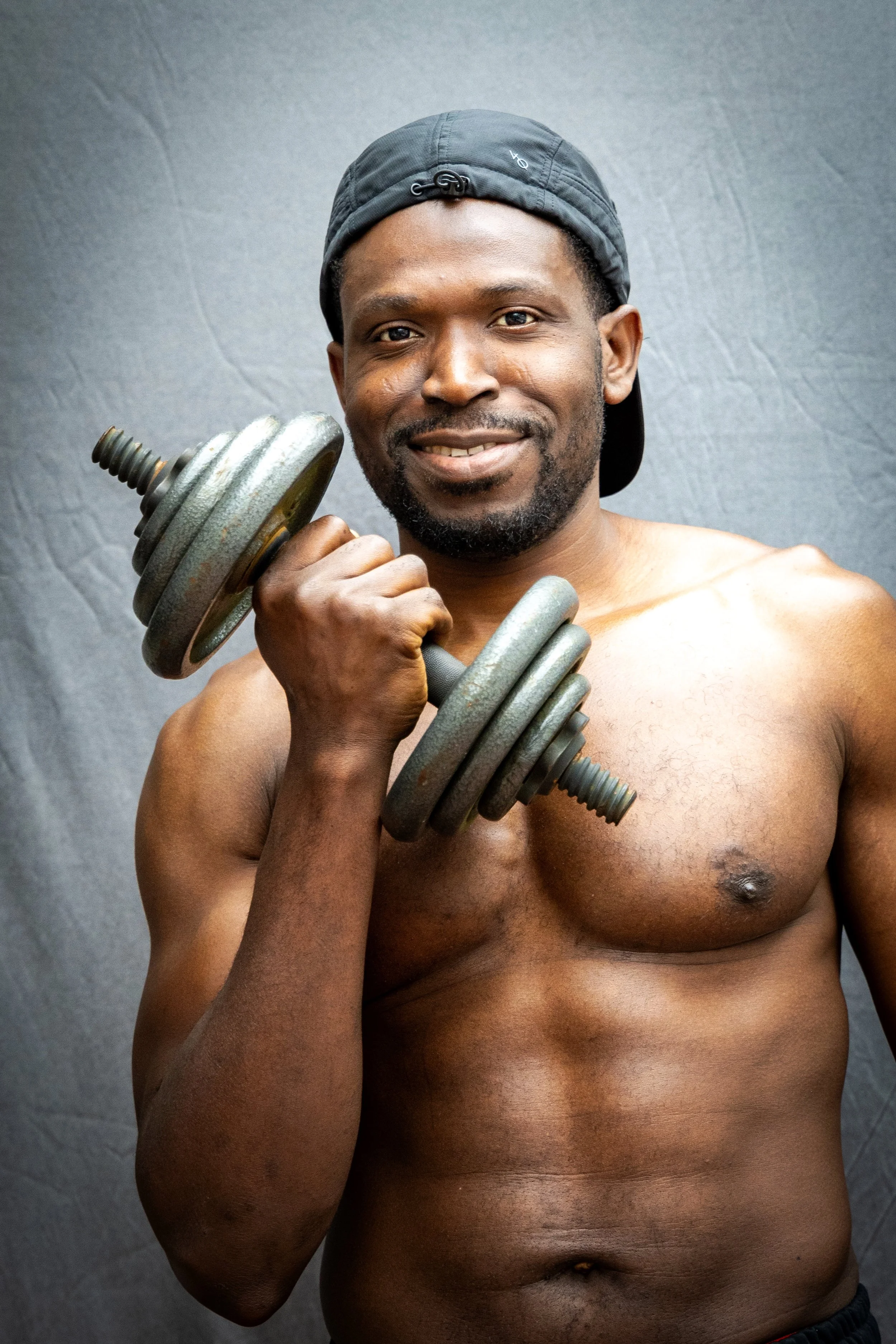 A shirtless man holds an adjustable dumbbell in one hand, standing against a plain gray background. He wears a backward black cap and has a friendly expression on his face.