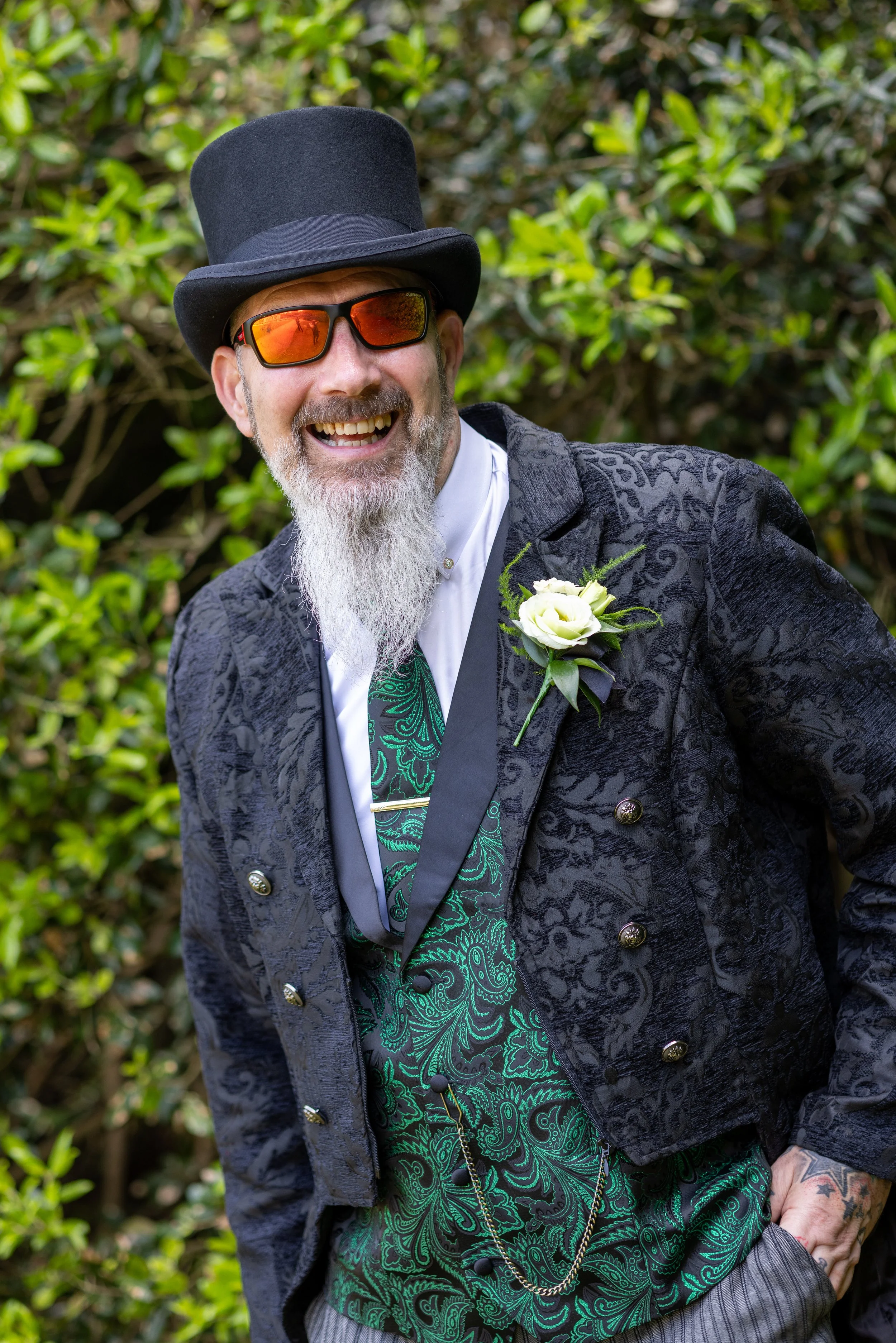 A man with a gray beard and mustache, wearing a black top hat and orange-tinted sunglasses, smiling widely. He is dressed in a black patterned blazer, a white shirt with a green paisley tie, and has a white flower boutonniere on his lapel. He is outd