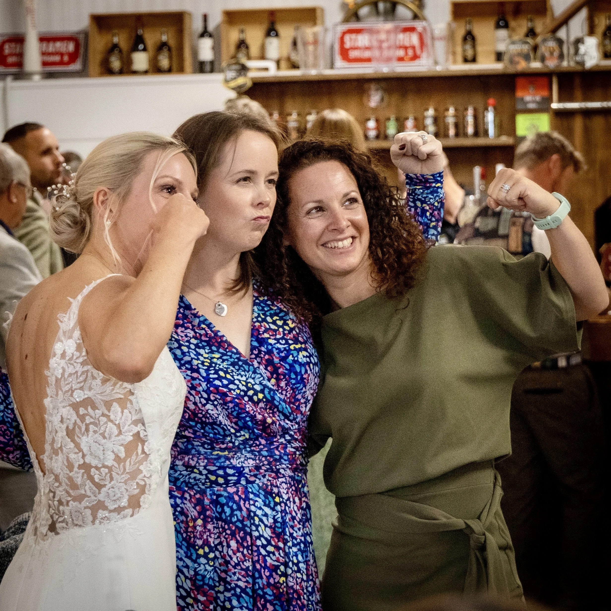 Three women in a restaurant, smiling and taking a selfie, with a background of shelves filled with bottles and signs.