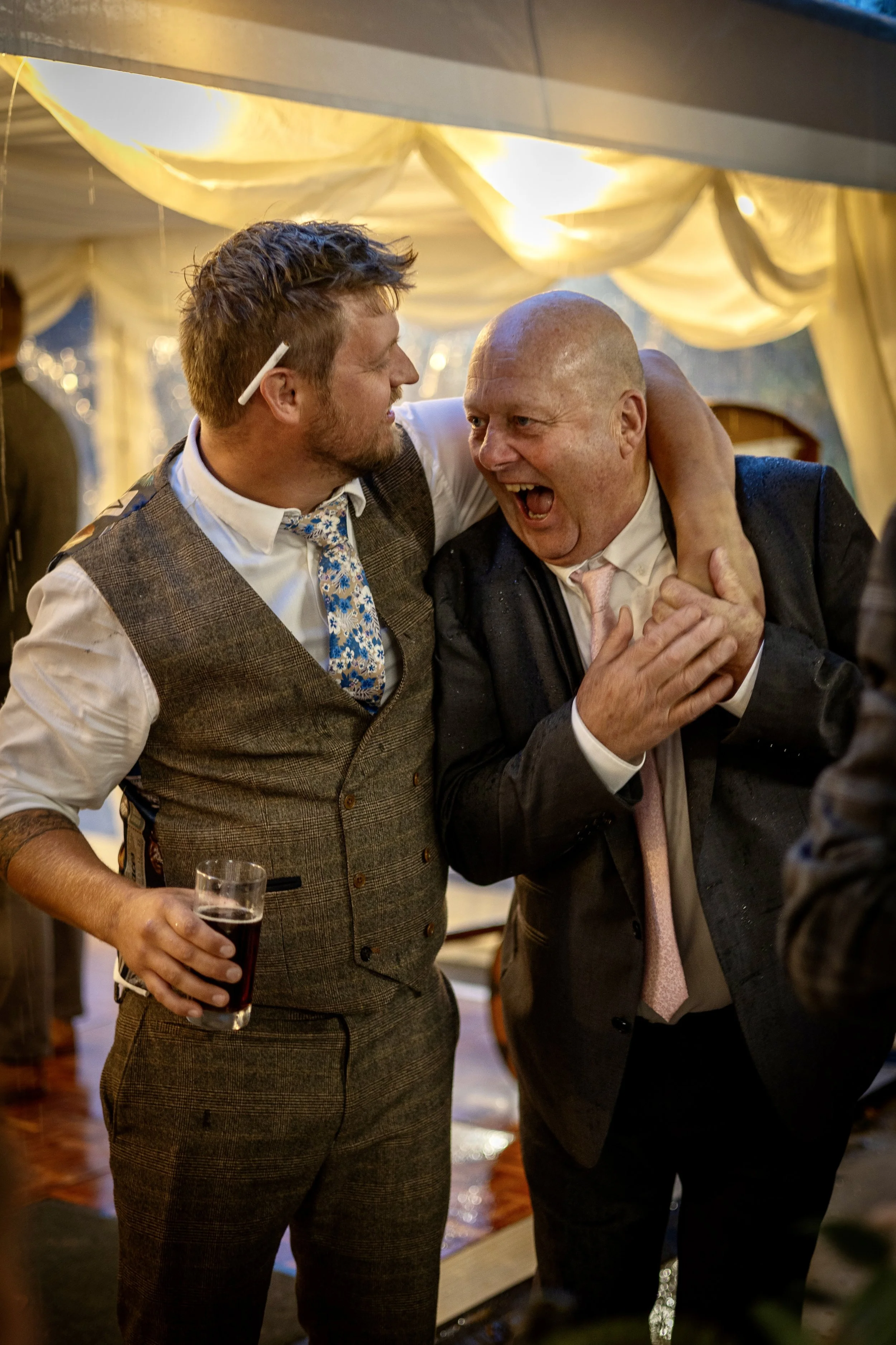 Two men sharing a joyful moment at a party or celebration, one holding a glass of dark beverage, with a decorated tent or marquee in the background.