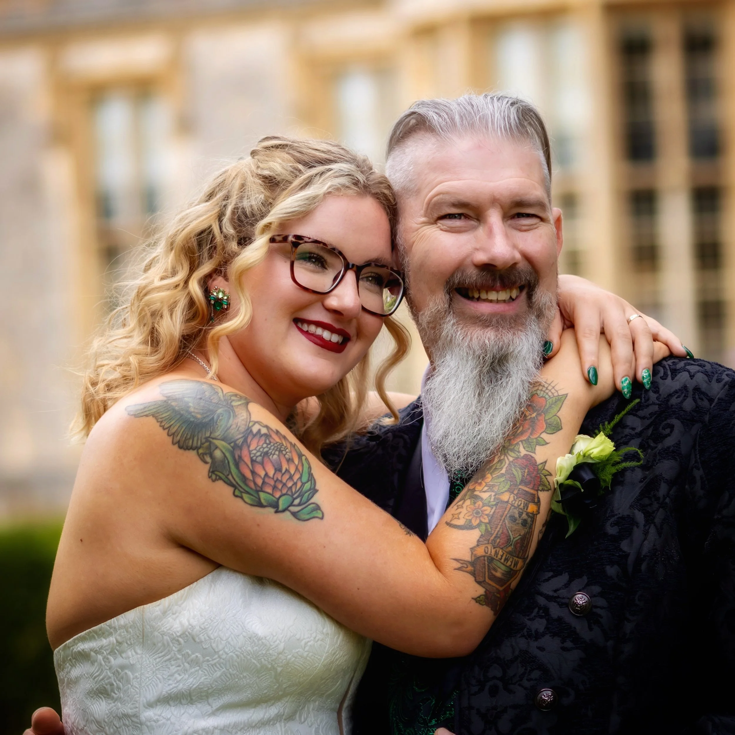 Close-up of a smiling couple embracing outdoors. The woman has curly blonde hair, glasses, and visible tattoos on her arm, and is wearing jewelry. The man has a grey beard, tattoos, and is wearing a dark patterned shirt. They are happy and appear to 